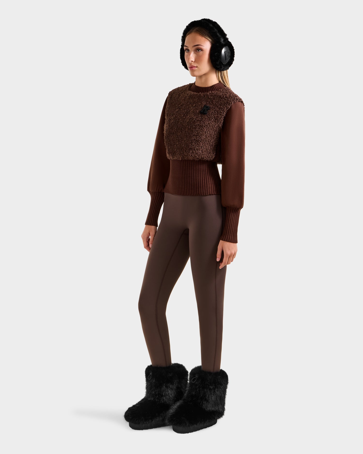 A woman wears the Rudsak FLORENCE - 8525546 Mocha top, paired with brown fitted pants, black fur boots, and black earmuffs—an après-ski essential look. She stands slightly turned against a plain light background.