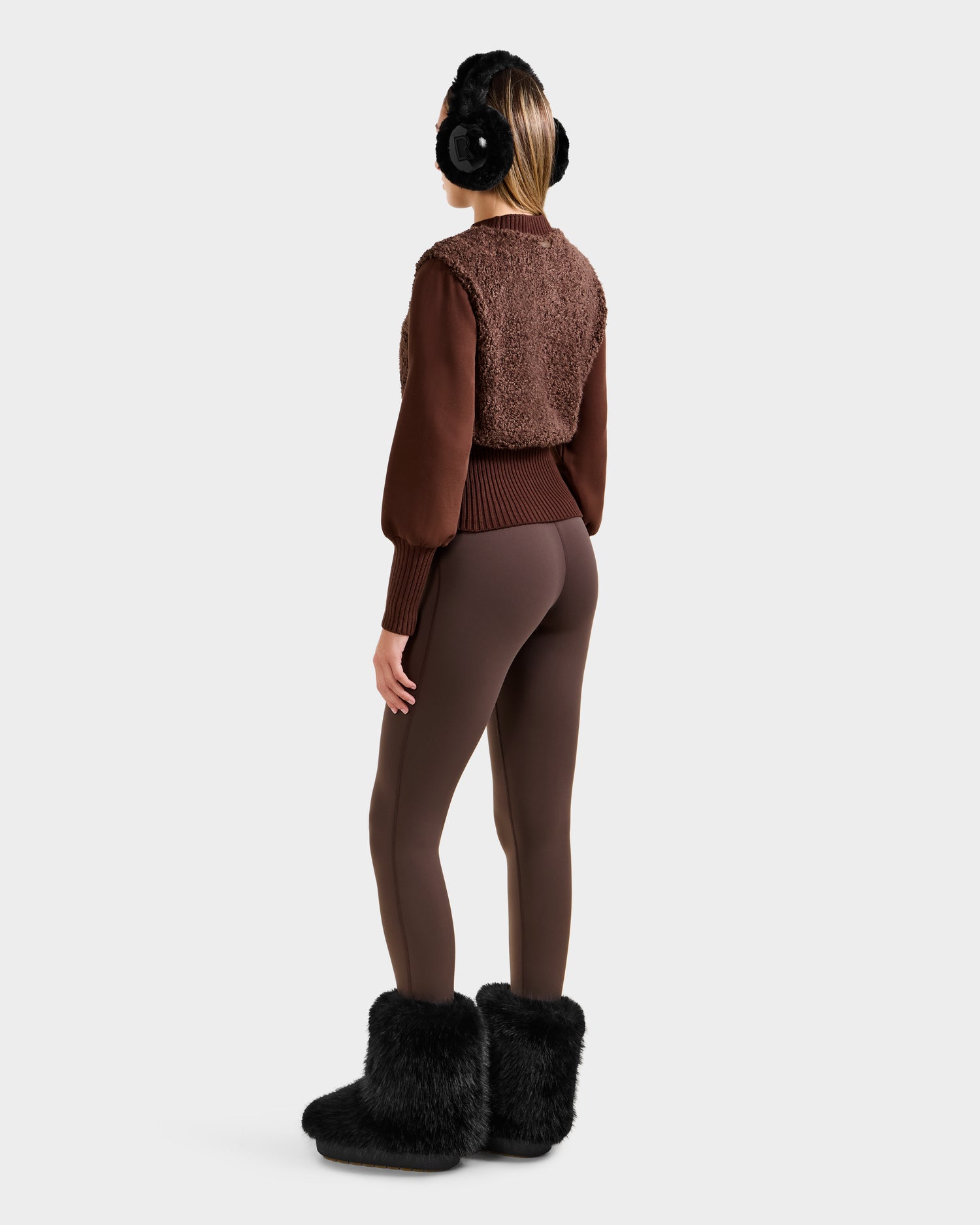 A woman faces away wearing Rudsaks FLORENCE - 8525546 Mocha faux fur vest over a brown sweater, paired with brown leggings and black earmuffs and furry boots, set against a plain light background—an après-ski essential.