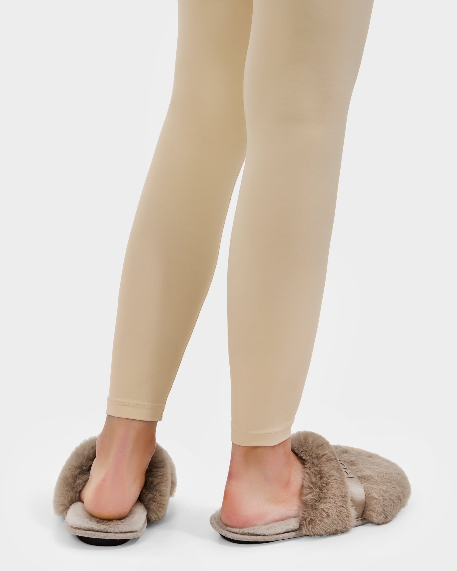 A person wearing beige leggings and Rudsak FLUFF - L223509 Almond faux-fur slippers with a cushioned insole stands on a white background, shown from the lower legs down—perfect for cozy downtime.