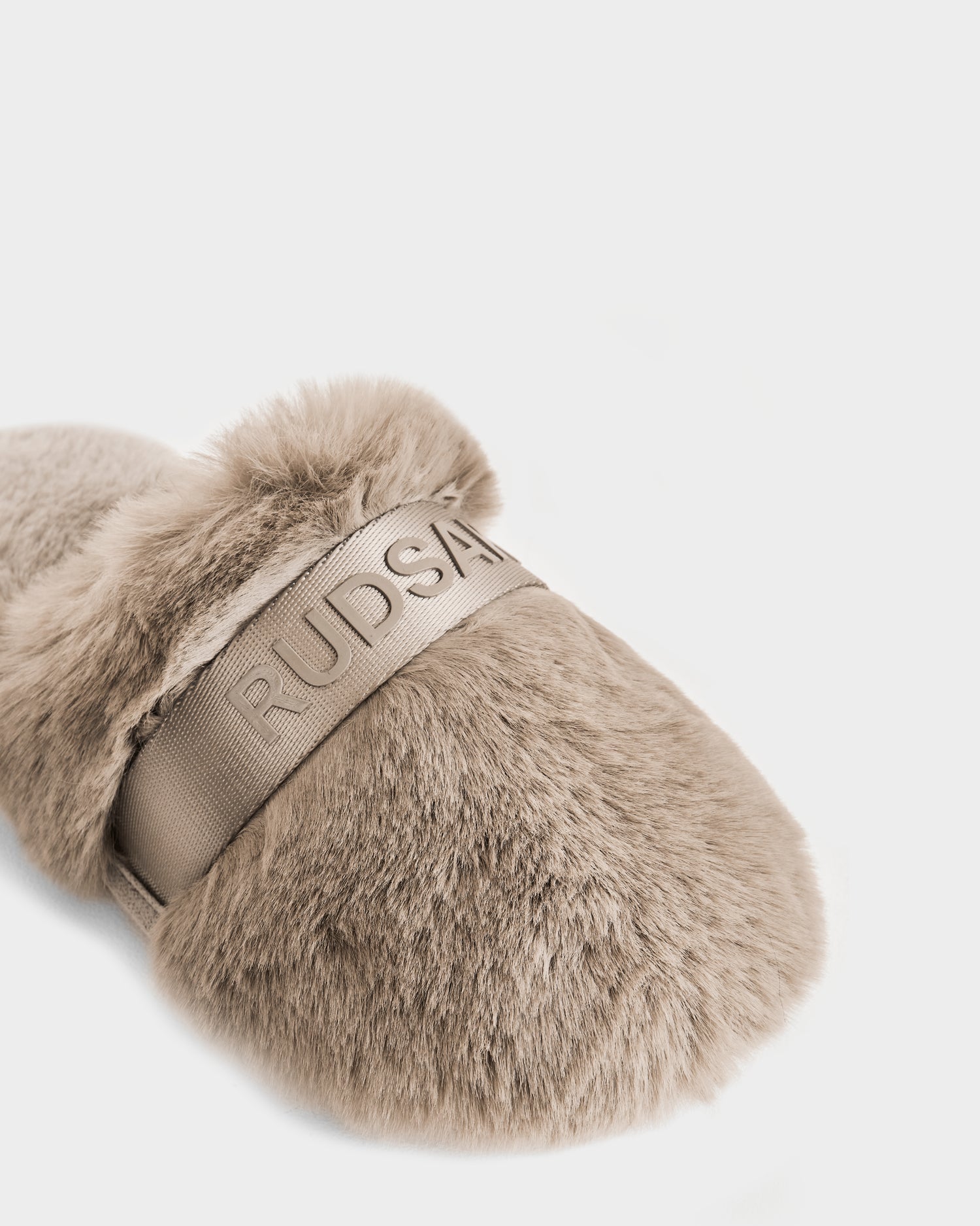 The FLUFF - L223509 Almond by Rudsak is a beige women’s slipper with thick faux fur and a wide strap embossed with RUDSAK branding, shown on a plain white background.