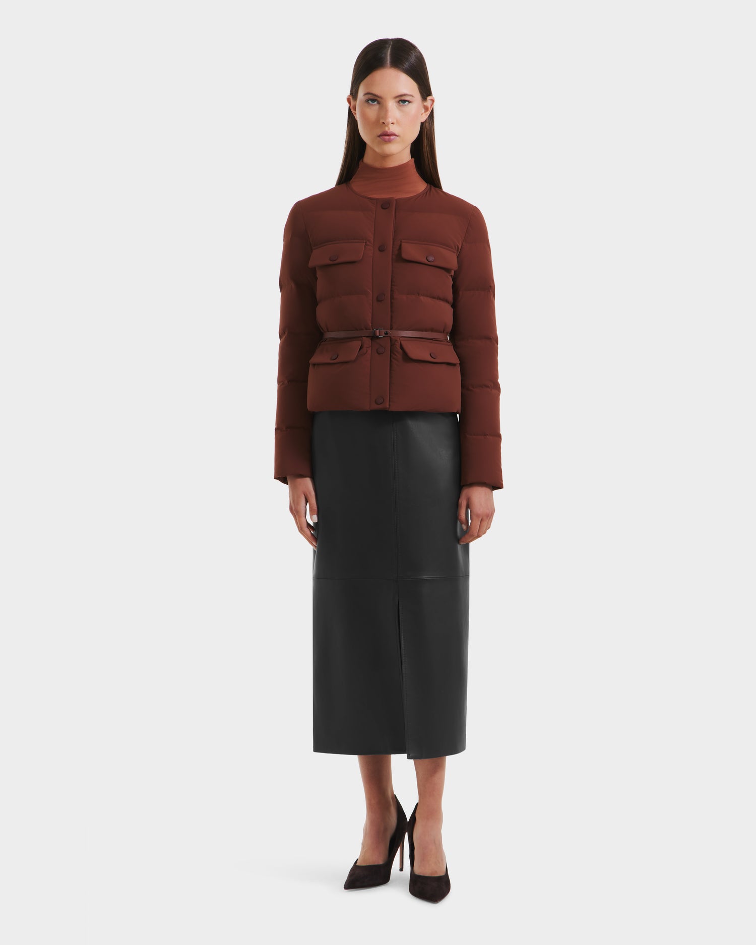A woman faces forward in the Rudsak FRANCESCA - 8125021 Rust fitted, dark red quilted puffer jacket with front pockets and a vegan leather belt, paired with a long black leather skirt and heels, set against a white background.