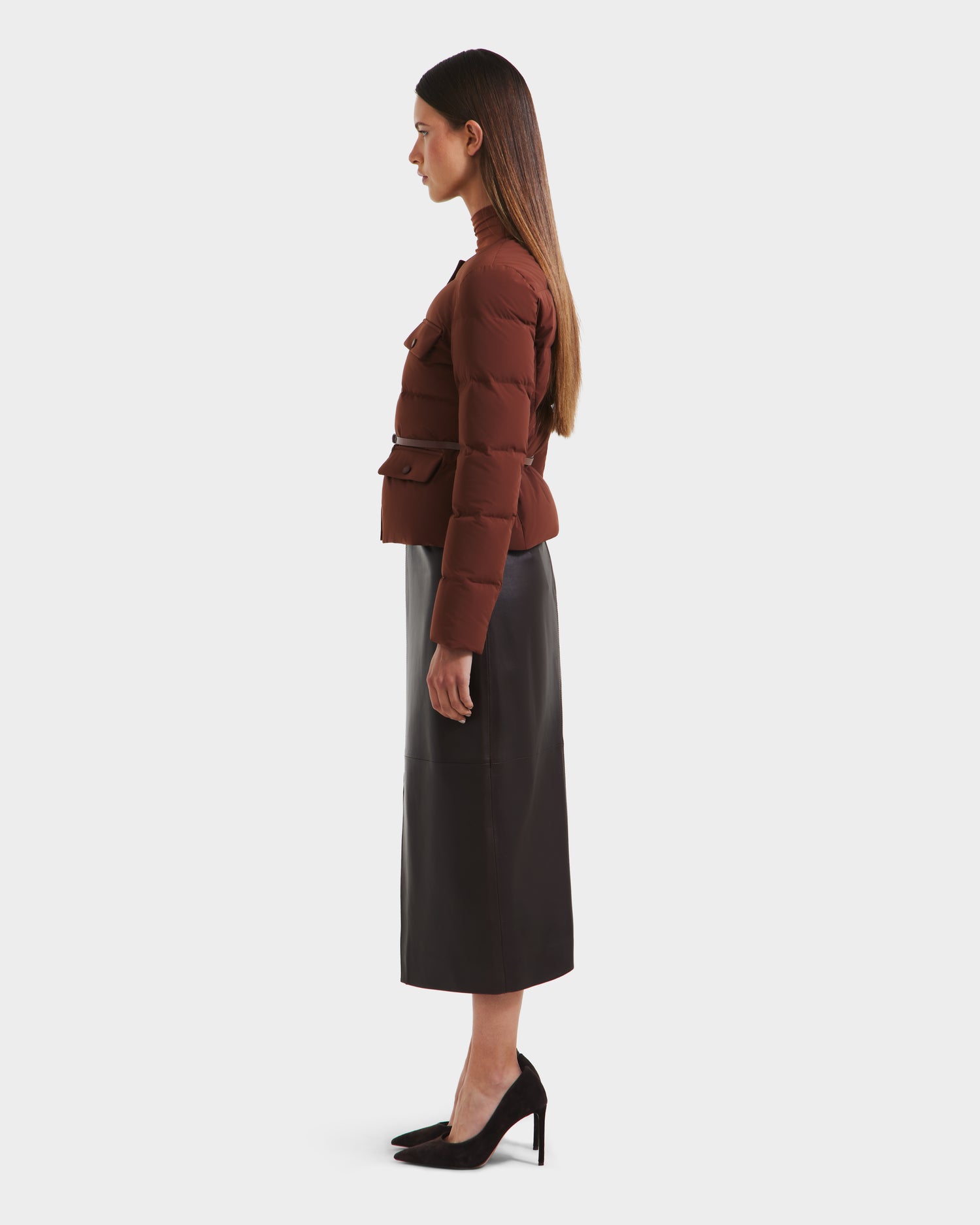 A woman stands in profile against a white background, wearing the Rudsak FRANCESCA - 8125021 Rust quilted puffer jacket, a black midi skirt with vegan leather belt, and black heels. Her long straight hair falls by her shoulders, arms relaxed at her sides.