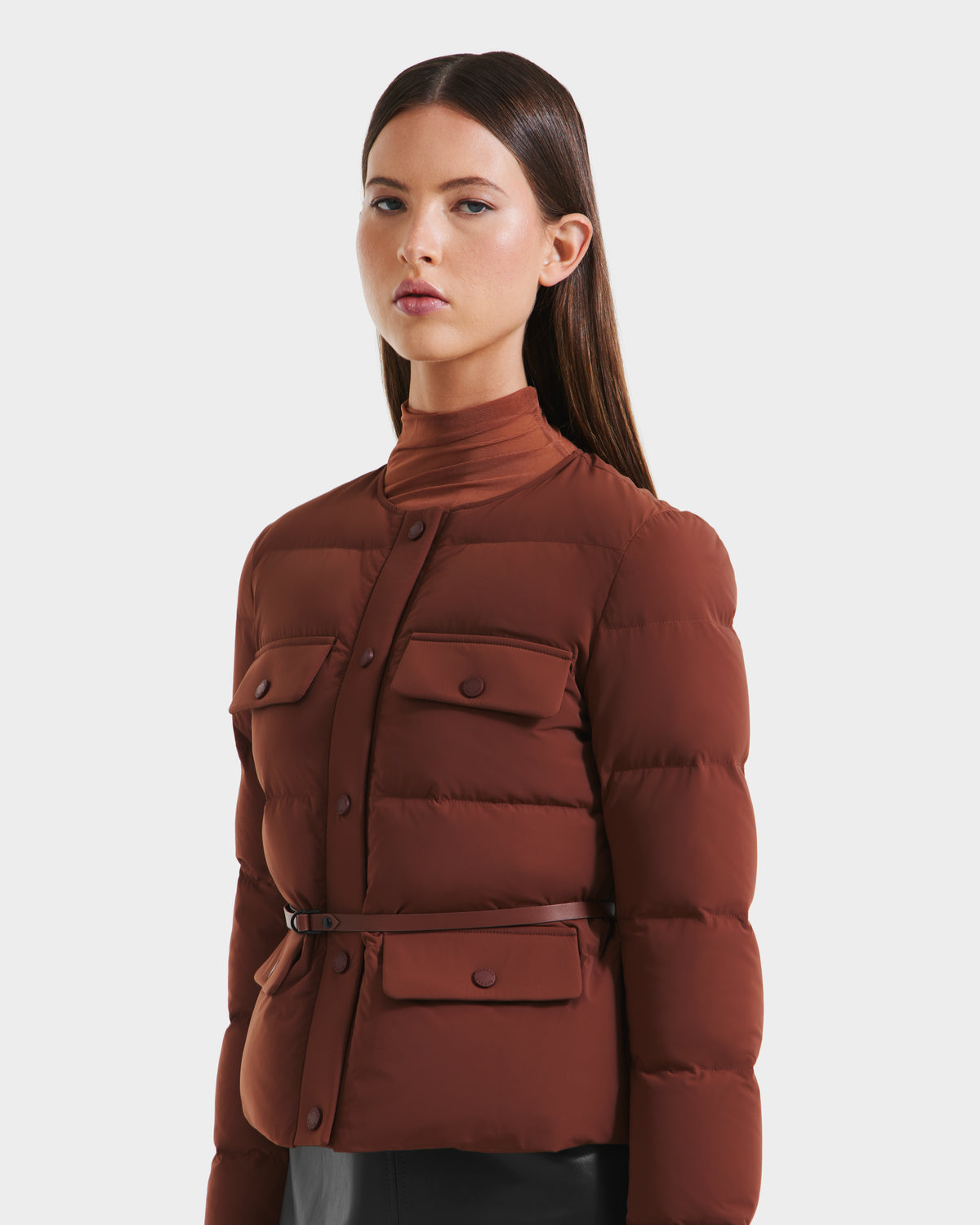A woman with straight brown hair models the Rudsak FRANCESCA - 8125021 Rust belted quilted puffer jacket, featuring flap pockets and a high-neck top, against a plain white background.