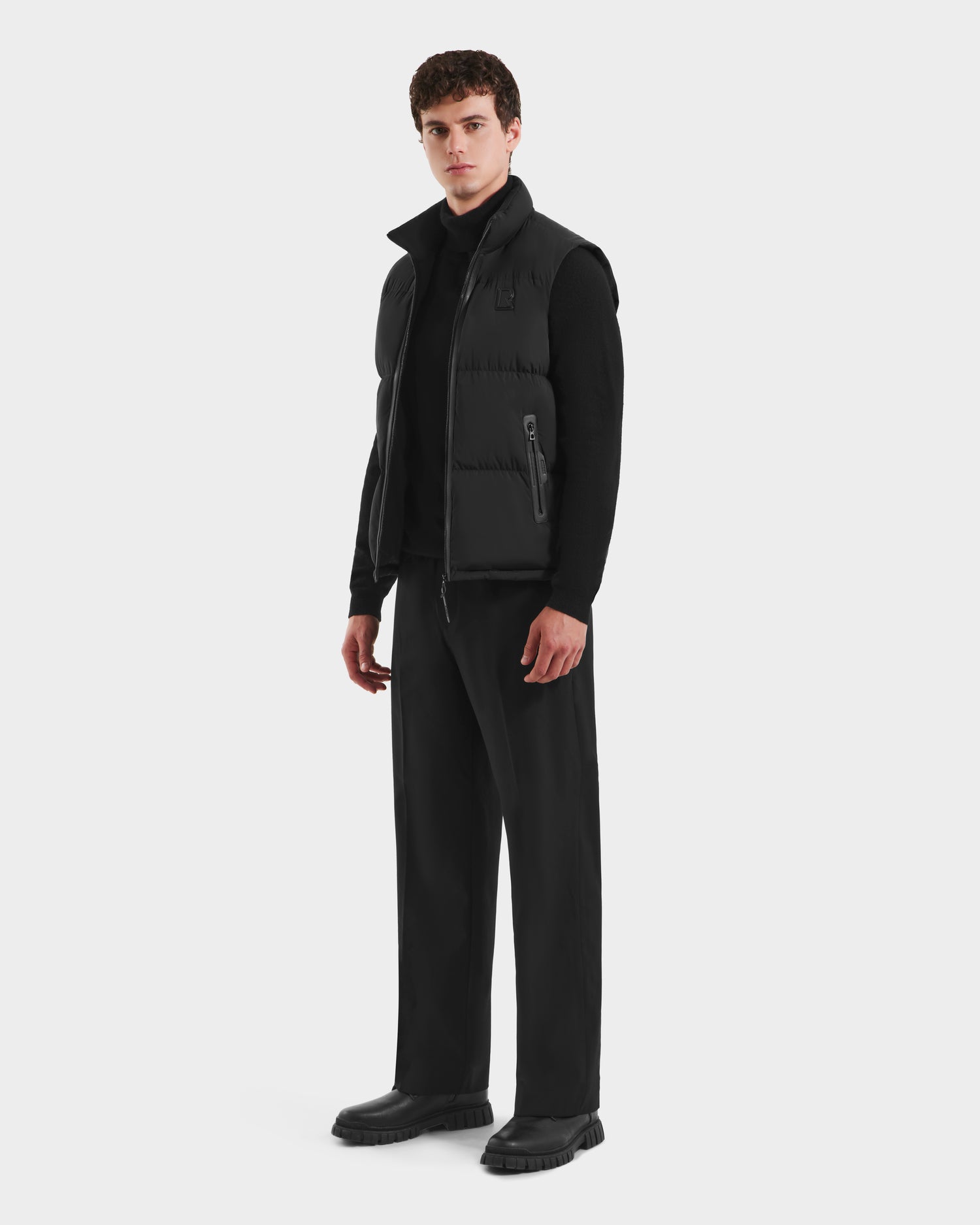 A person stands facing forward, wearing the Rudsak FREDDY - 6125571 Black men’s puffer vest over a black long-sleeve shirt, black pants, and black shoes—perfect for lightweight layering against a plain white background.