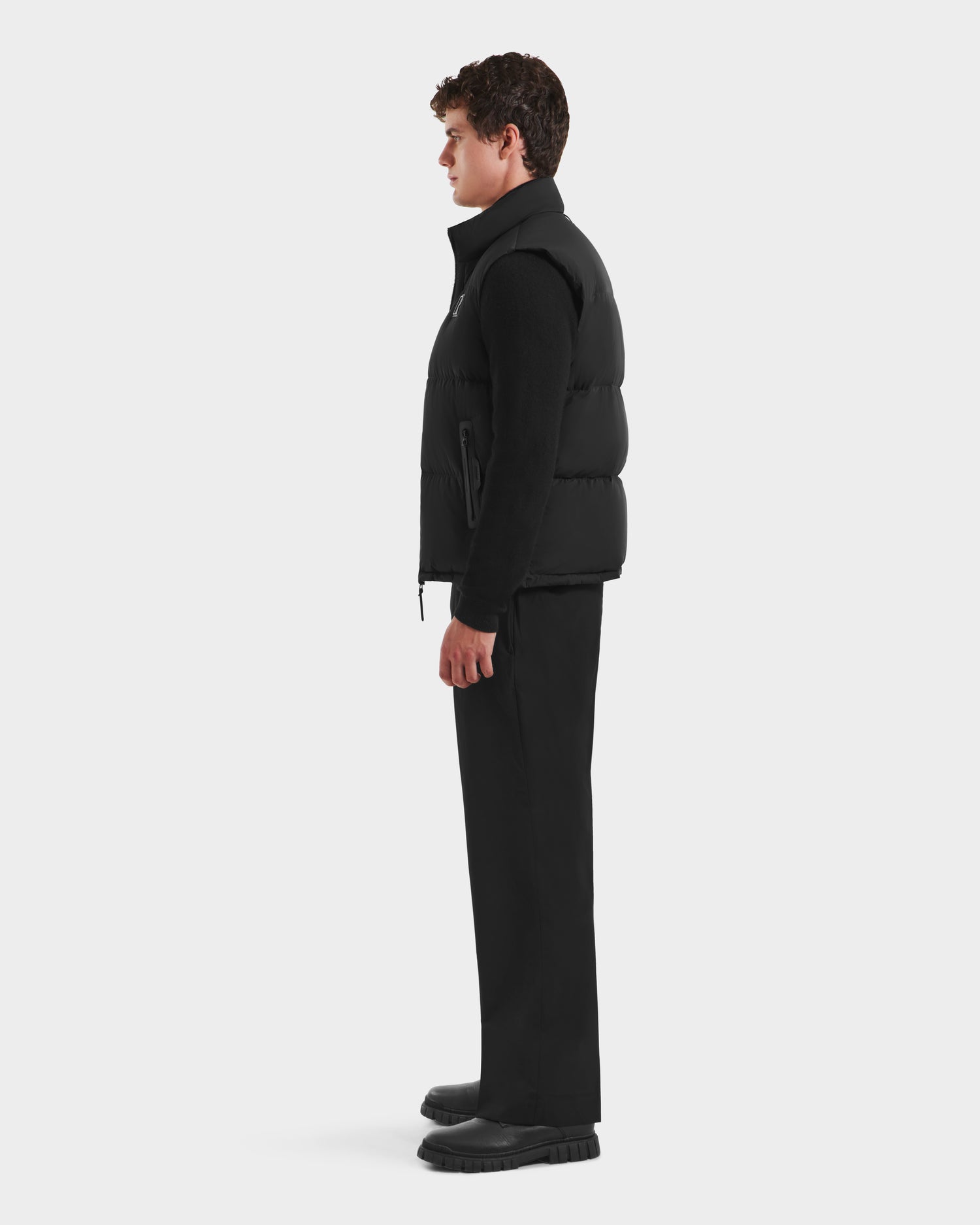 A person stands in profile against a plain background, wearing the FREDDY - 6125571 Black men’s puffer vest by Rudsak over a black long-sleeve top, paired with black pants and shoes—ideal for lightweight layering.