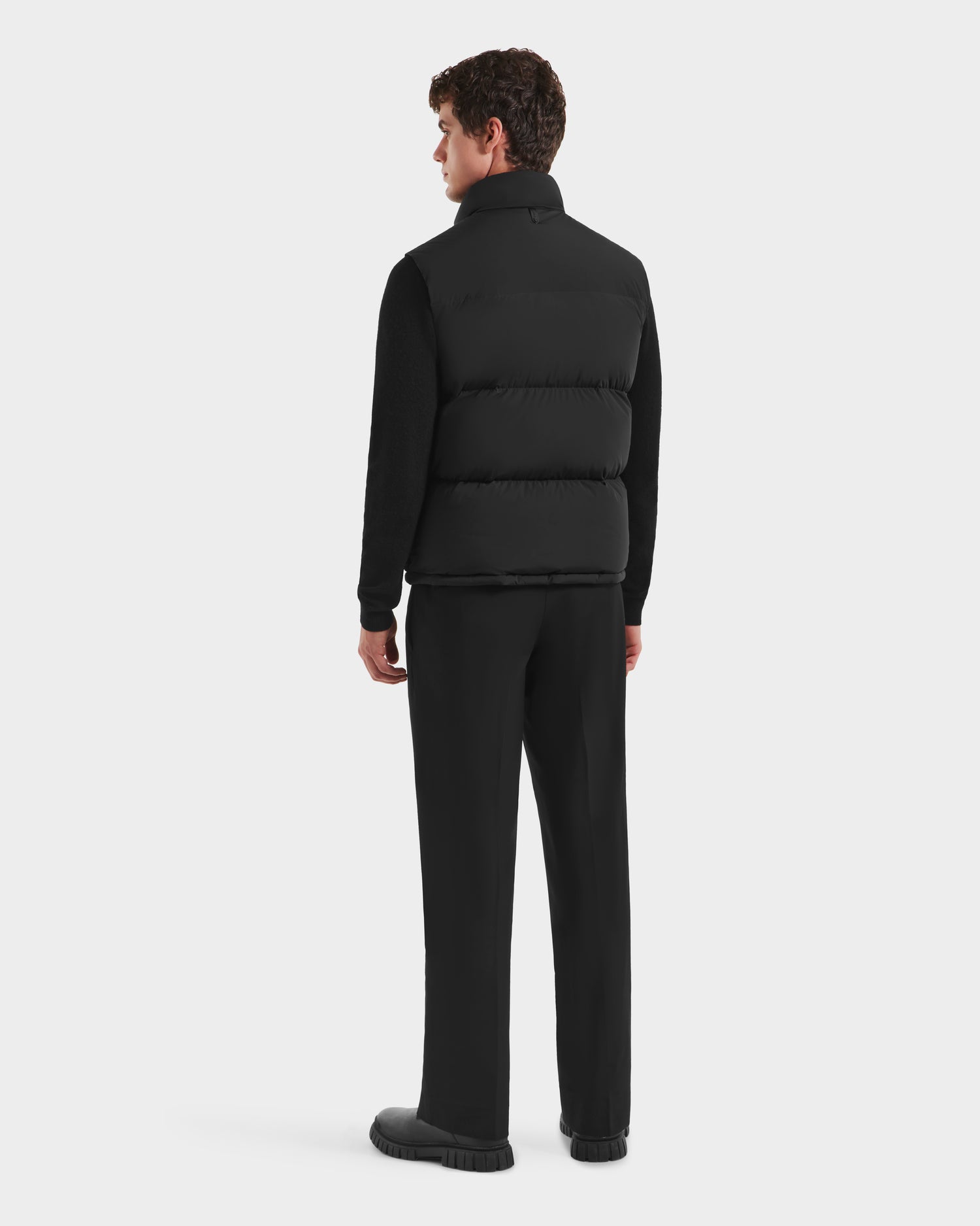 A person with short, curly brown hair faces away, wearing the Rudsak FREDDY - 6125571 Black men’s puffer vest over black long sleeves, pants, and shoes against a plain white background.