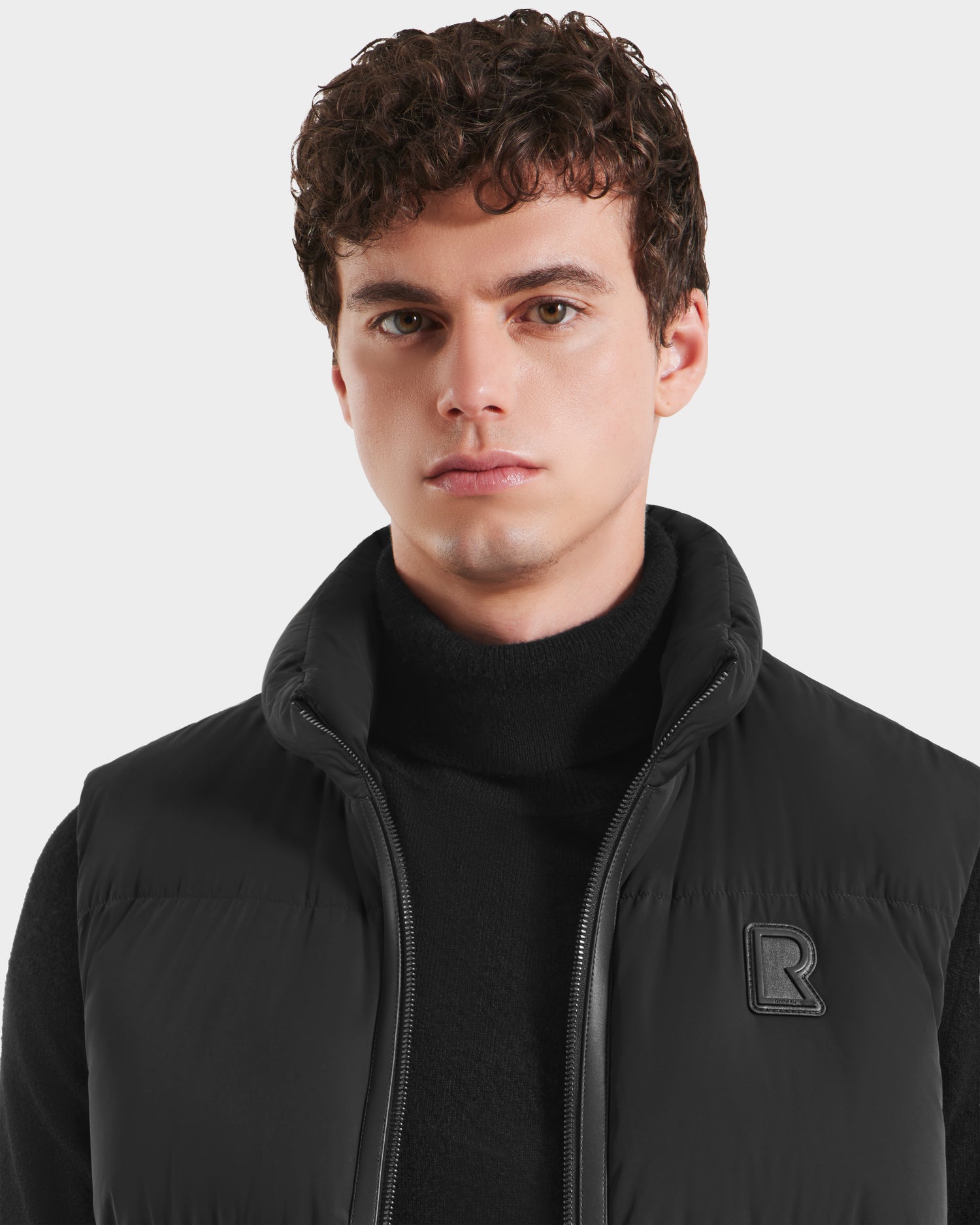 A young man with short curly brown hair wears a black turtleneck and the Rudsak FREDDY - 6125571 Black men’s puffer vest with a B logo, ideal for lightweight layering, standing against a plain light background.
