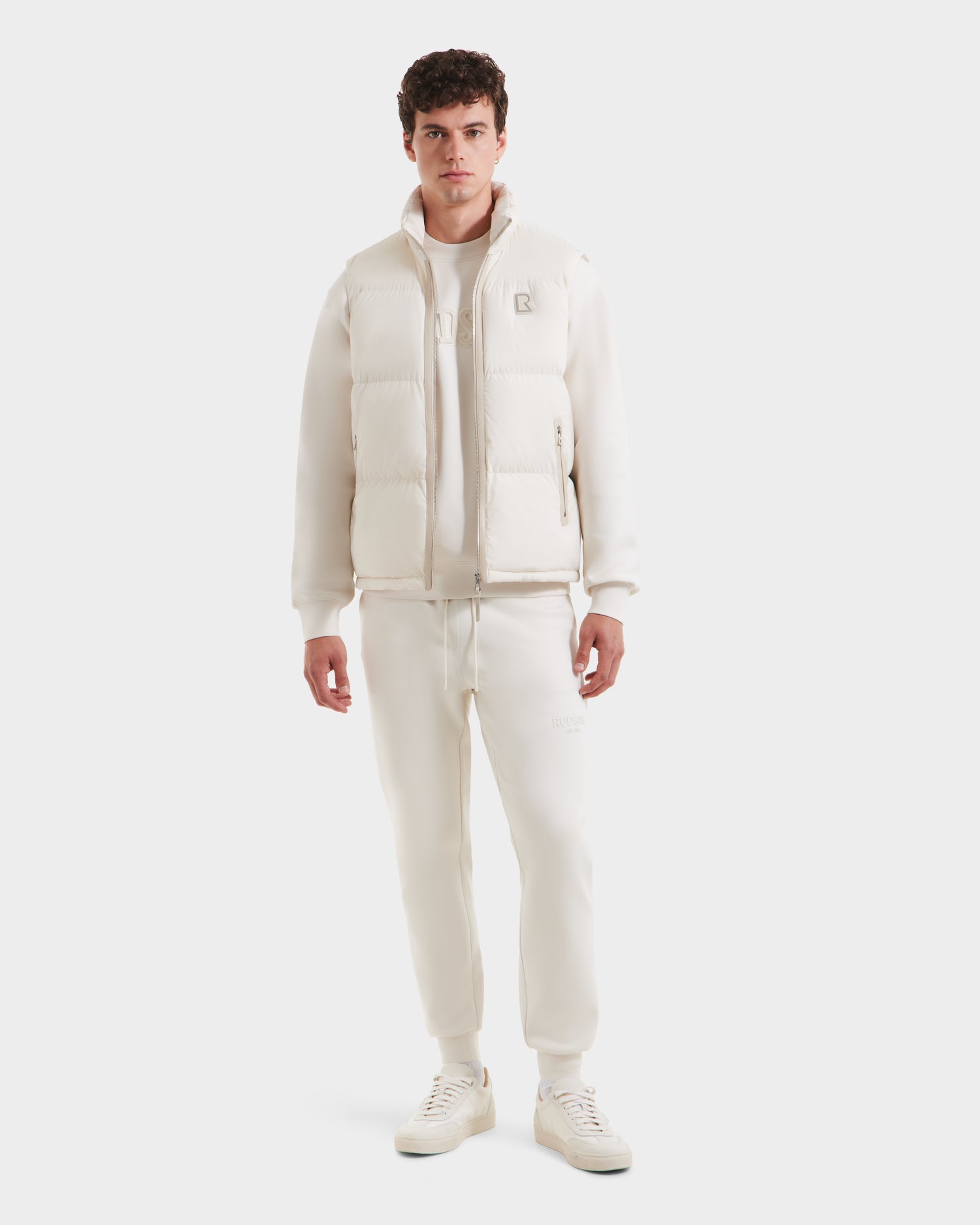 A man faces forward against a plain white background, dressed in the Rudsak FREDDY - 6125571 Vanilla men’s puffer vest, paired with a sweatshirt, sweatpants, and sneakers for a sleek all-white layered look.