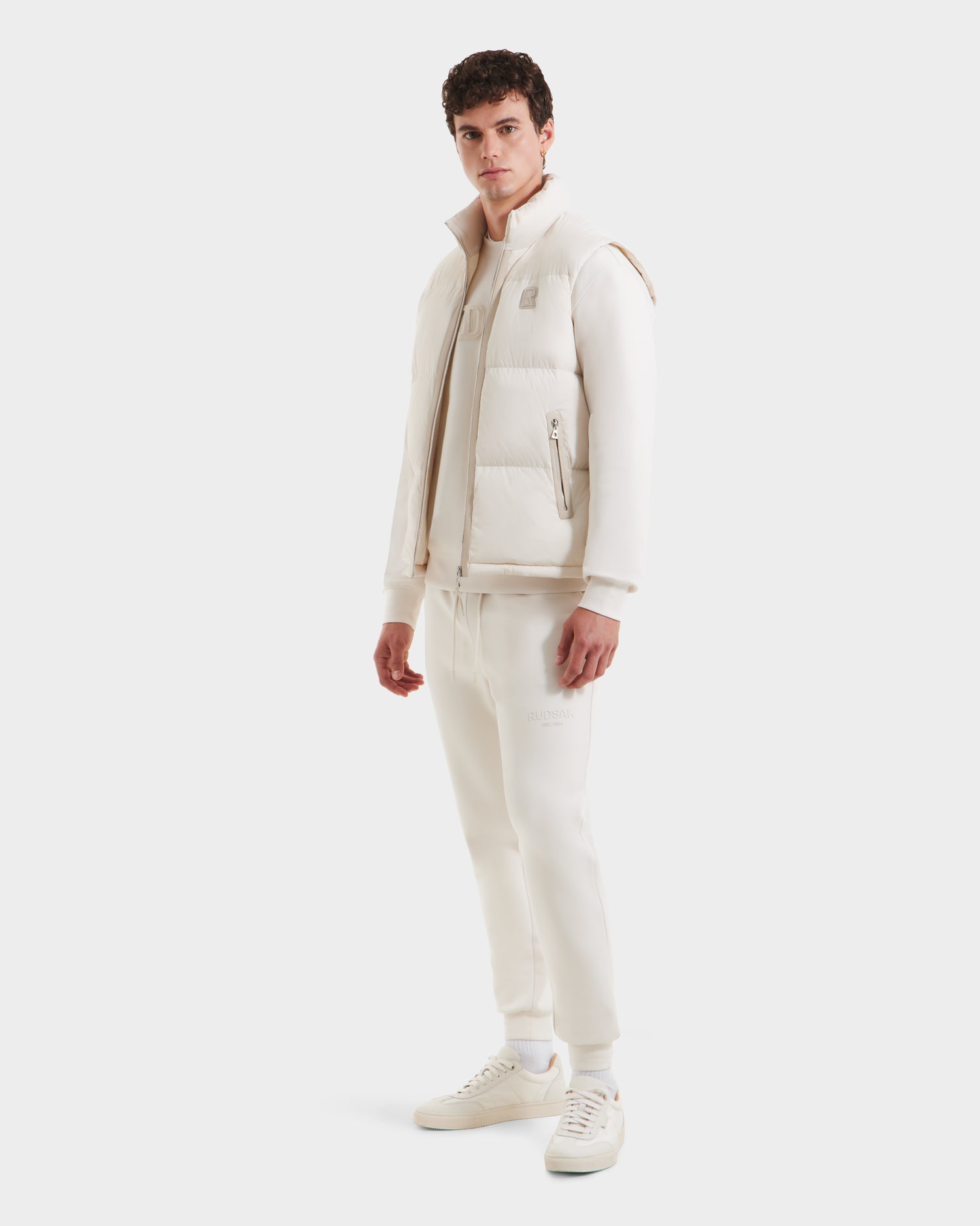 A man faces forward against a plain white background, wearing the Rudsak FREDDY - 6125571 Vanilla men’s puffer vest layered over a sweatshirt, with sweatpants and sneakers for a clean, casual look.