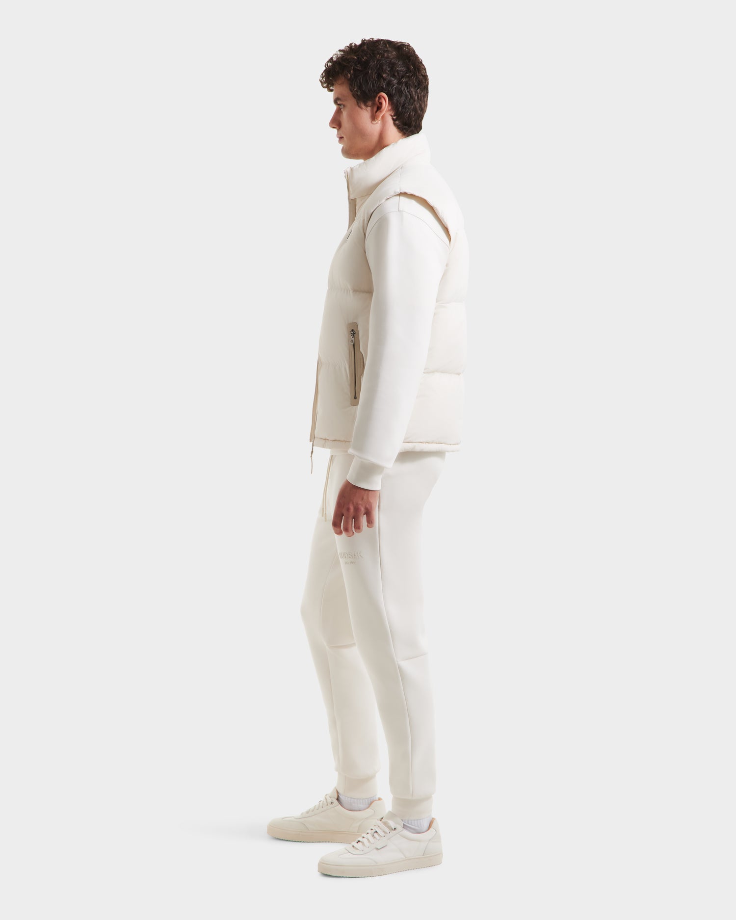 A man stands in profile against a white background, wearing the Rudsak FREDDY - 6125571 Vanilla puffer vest, ideal for lightweight layering over a long-sleeve shirt, paired with matching pants and white sneakers.