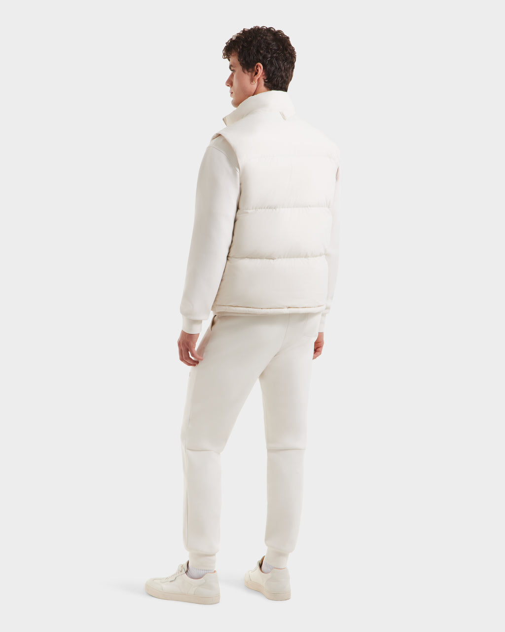 Facing away, a man wears the Rudsak FREDDY - 6125571 Vanilla men’s puffer vest. Paired with an off-white sweatshirt, sweatpants, and sneakers, his look is perfect for lightweight layering against a softly colored backdrop.