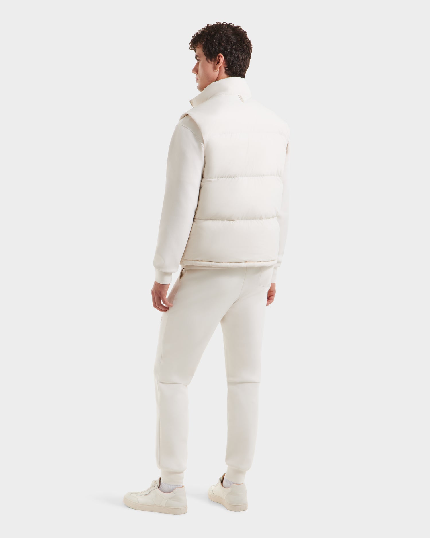 Facing away, a man wears the Rudsak FREDDY - 6125571 Vanilla men’s puffer vest. Paired with an off-white sweatshirt, sweatpants, and sneakers, his look is perfect for lightweight layering against a softly colored backdrop.