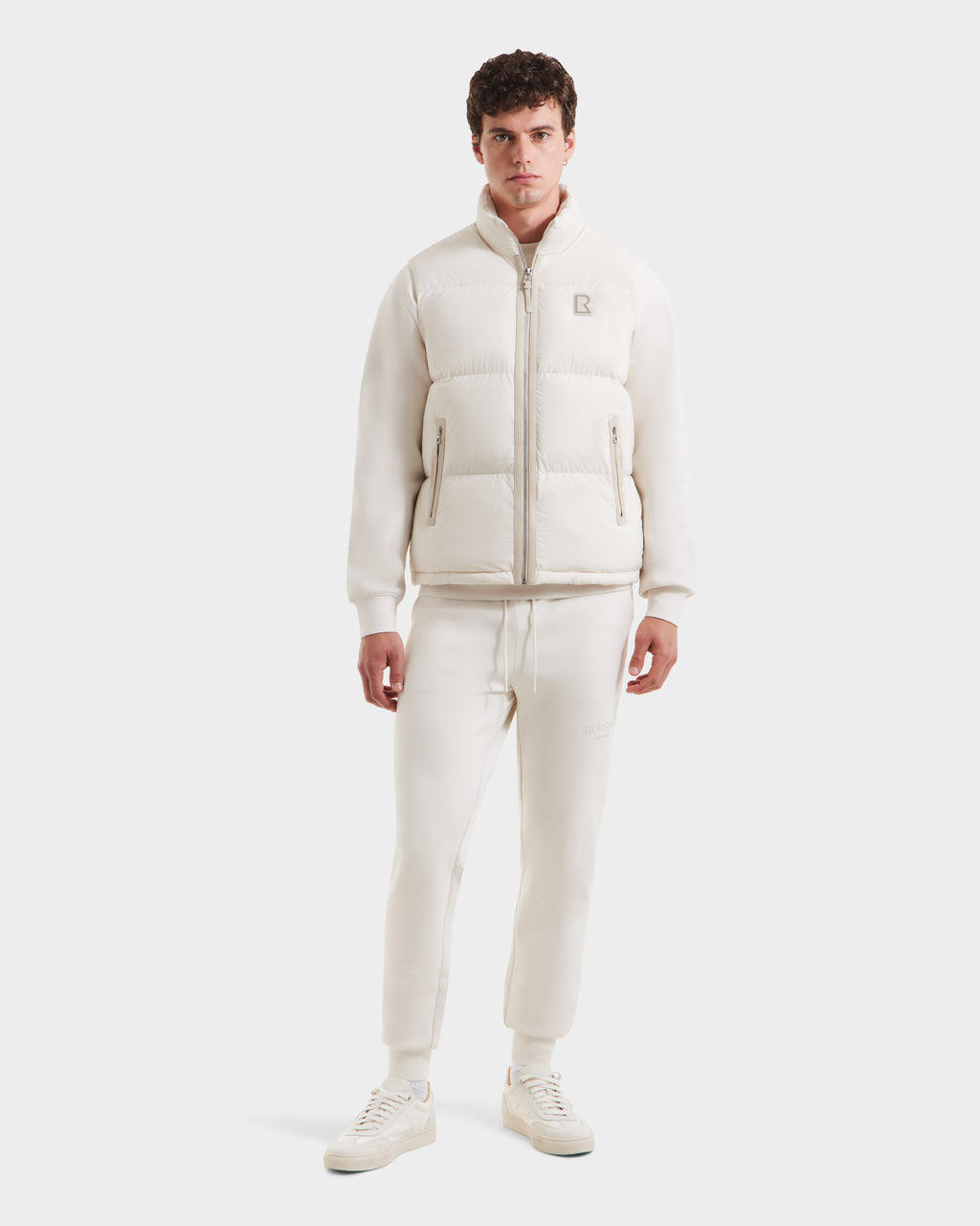 A man faces forward against a white background, wearing Rudsak’s FREDDY - 6125571 Vanilla jogger-style pants and sneakers—lightweight layering ideal for men’s puffer vest season.
