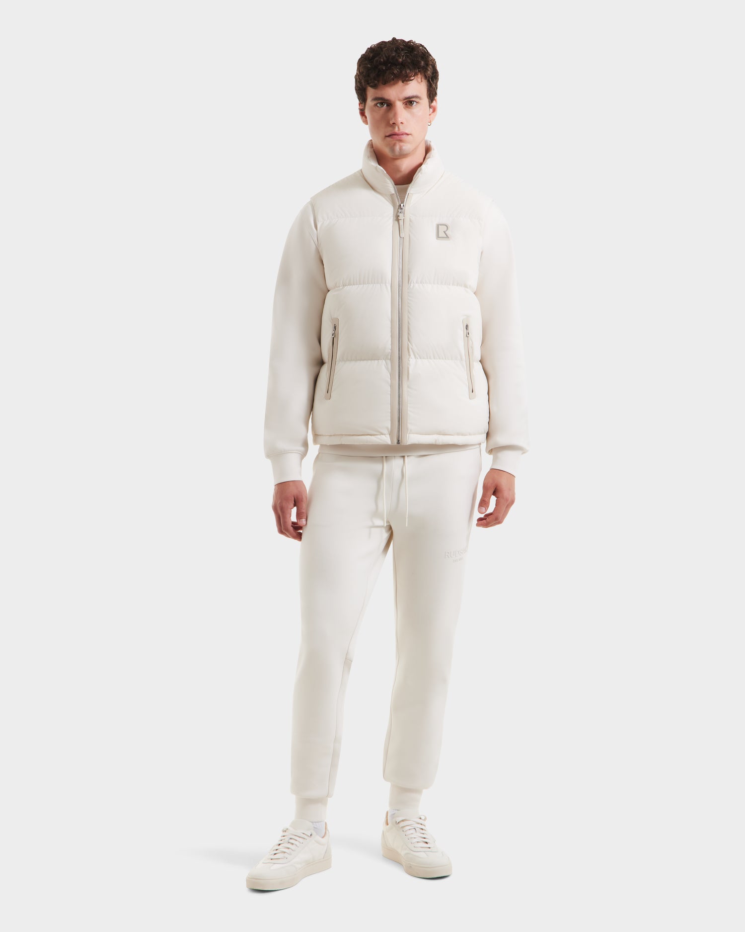 A man faces forward against a white background, wearing Rudsak’s FREDDY - 6125571 Vanilla jogger-style pants and sneakers—lightweight layering ideal for men’s puffer vest season.