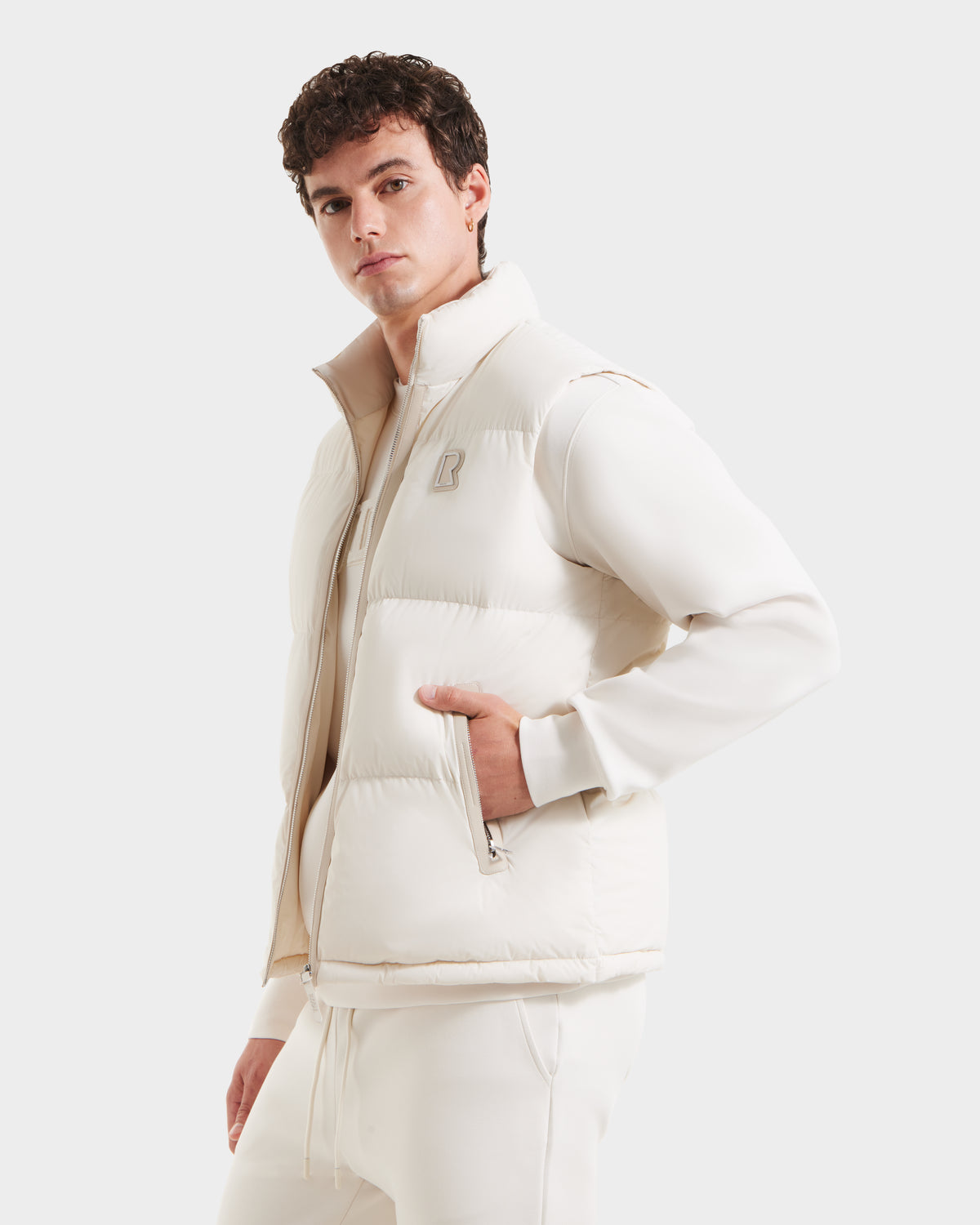 A young man models the Rudsak FREDDY - 6125571 Vanilla premium down puffer vest over a matching sweatshirt and pants, standing confidently with one hand in his pocket—a stylish, lightweight layering option.