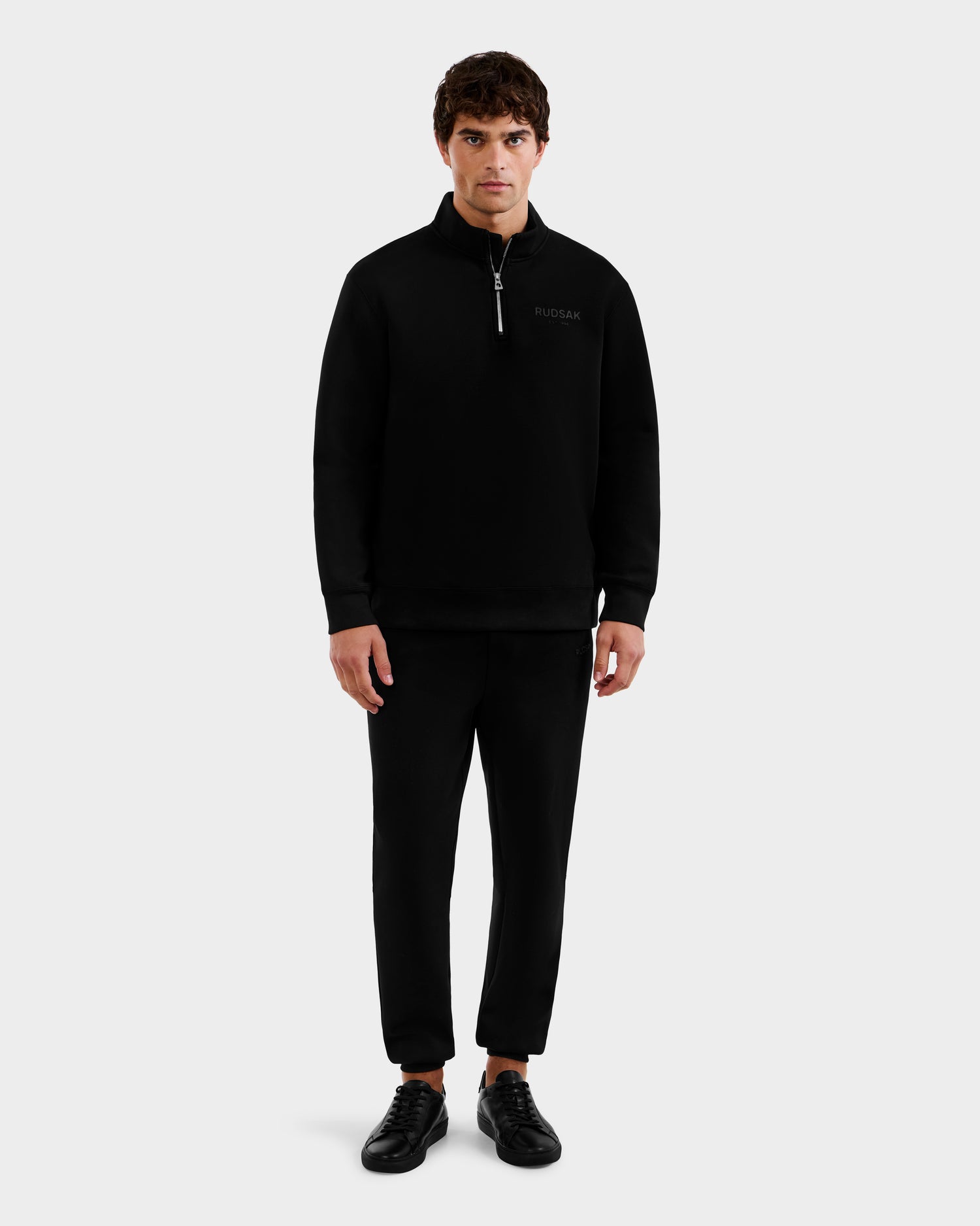 A man faces forward against a light gray background, wearing the Rudsak FREMONT - 9525000 Black M mock neck cotton sweater with matching black pants and black sneakers.