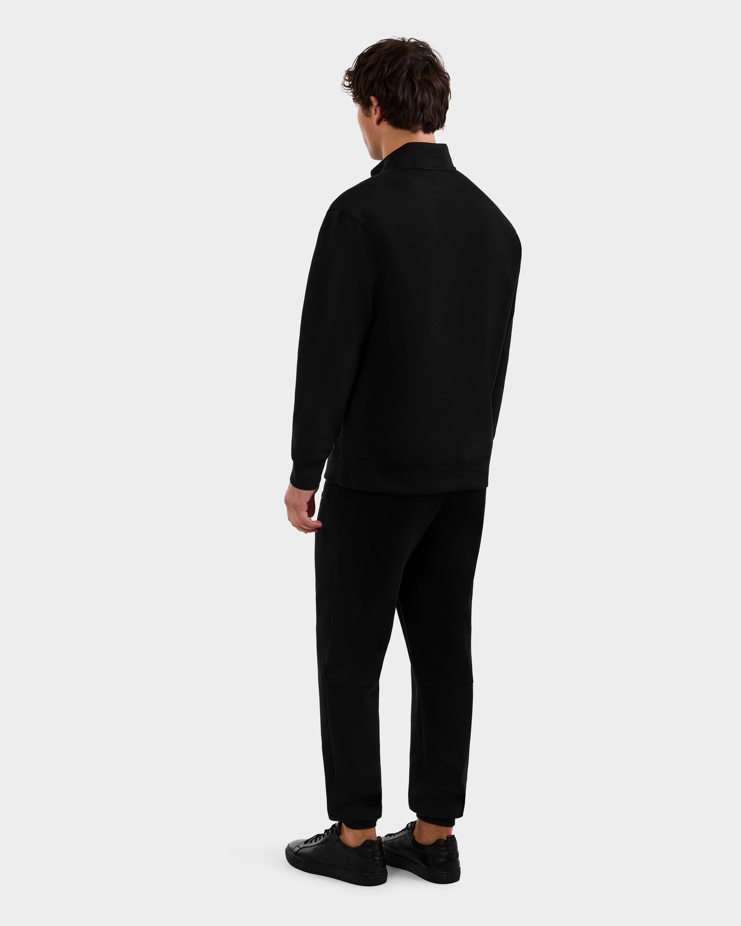 A man faces away from the camera, wearing the Rudsak FREMONT - 9525000 Black M mock neck sweater with black pants and sneakers, against a plain light gray background.