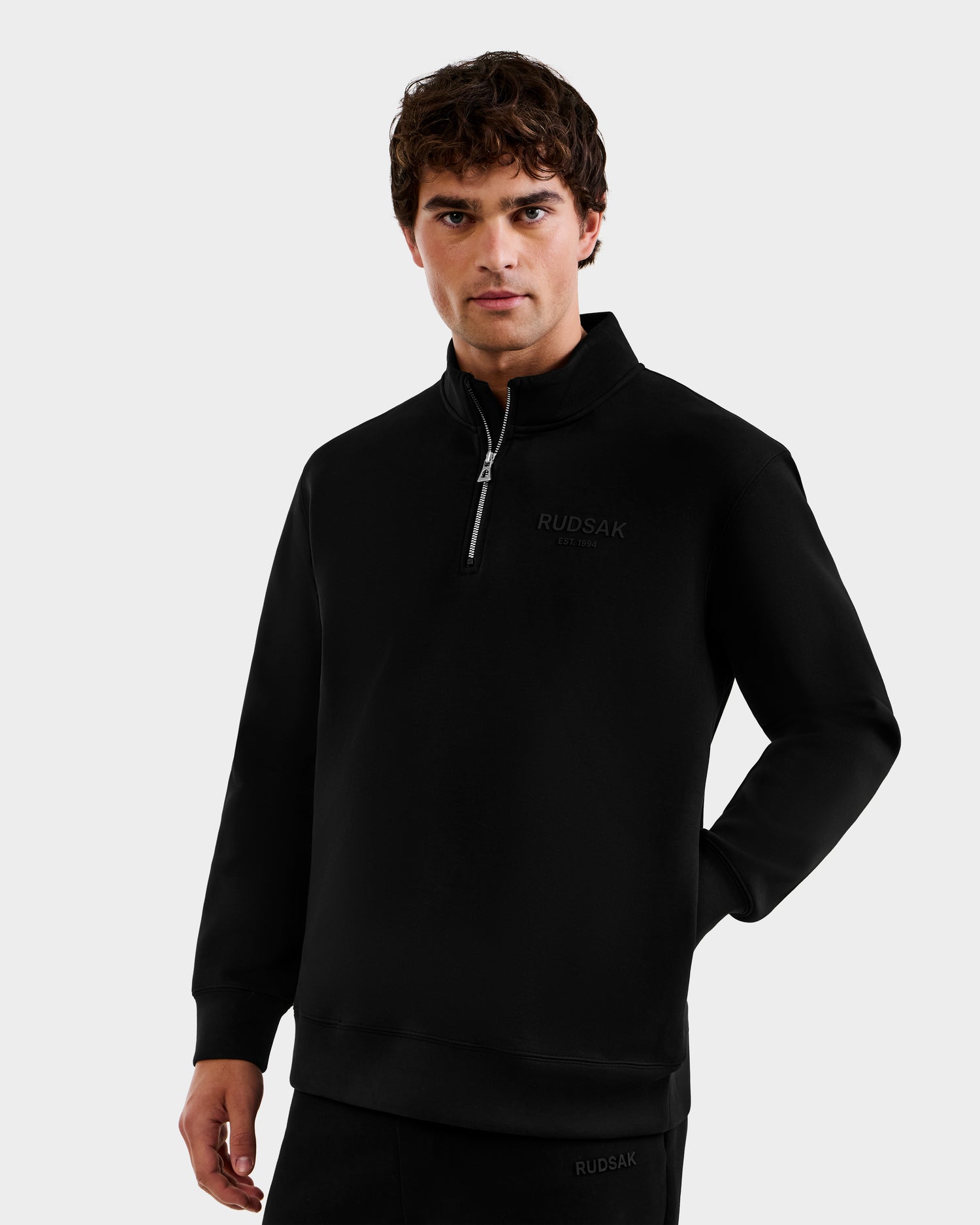 A man in a Rudsak FREMONT - 9525000 Black M quarter-zip pullover with "RUDSAK" on the chest stands against a plain light background, one hand by his side and the other arm resting on his hip.