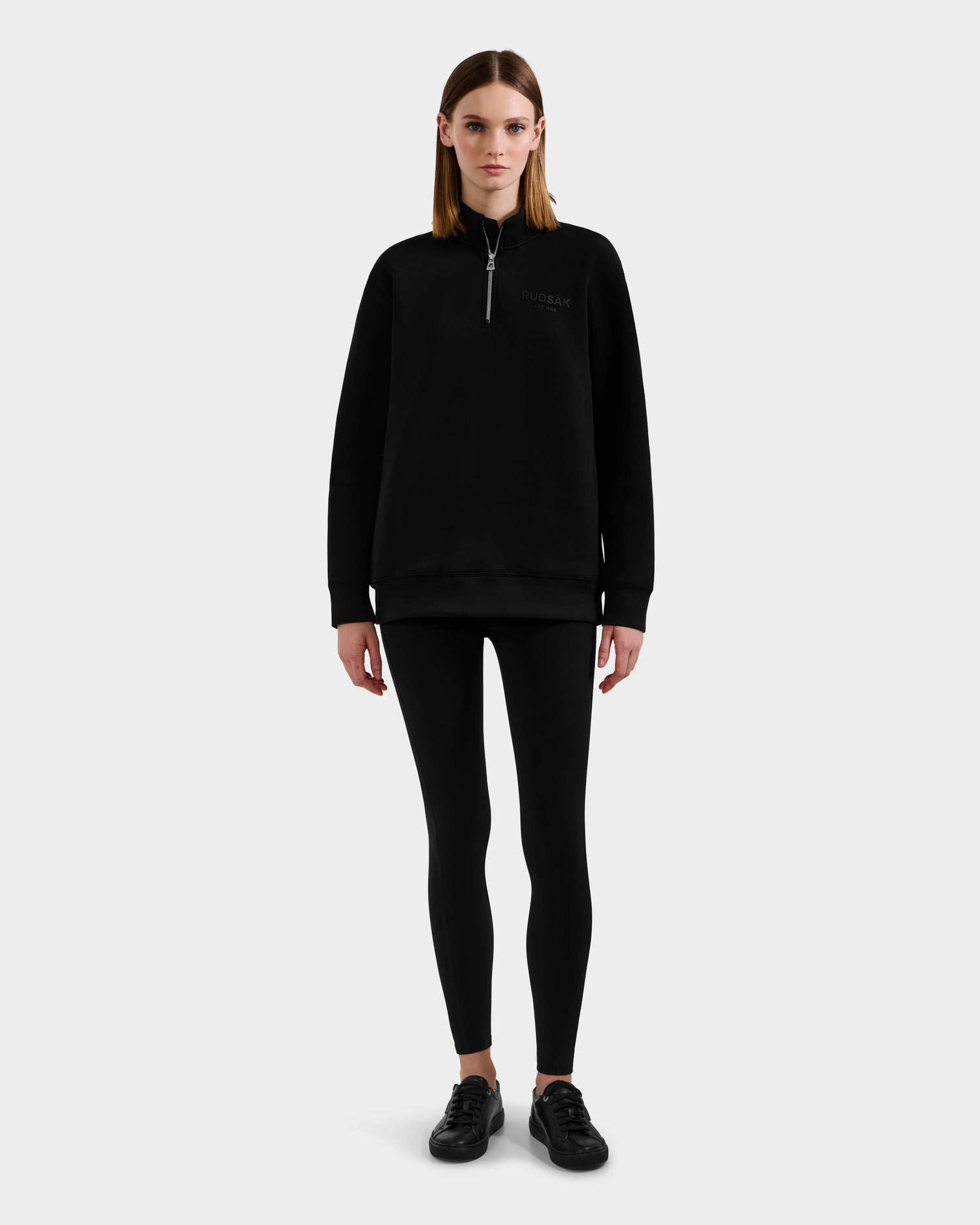 A woman faces forward in a Rudsak FREMONT - 9525000 Black W quarter-zip sweatshirt, paired with black leggings and sneakers, set against a plain light gray background.
