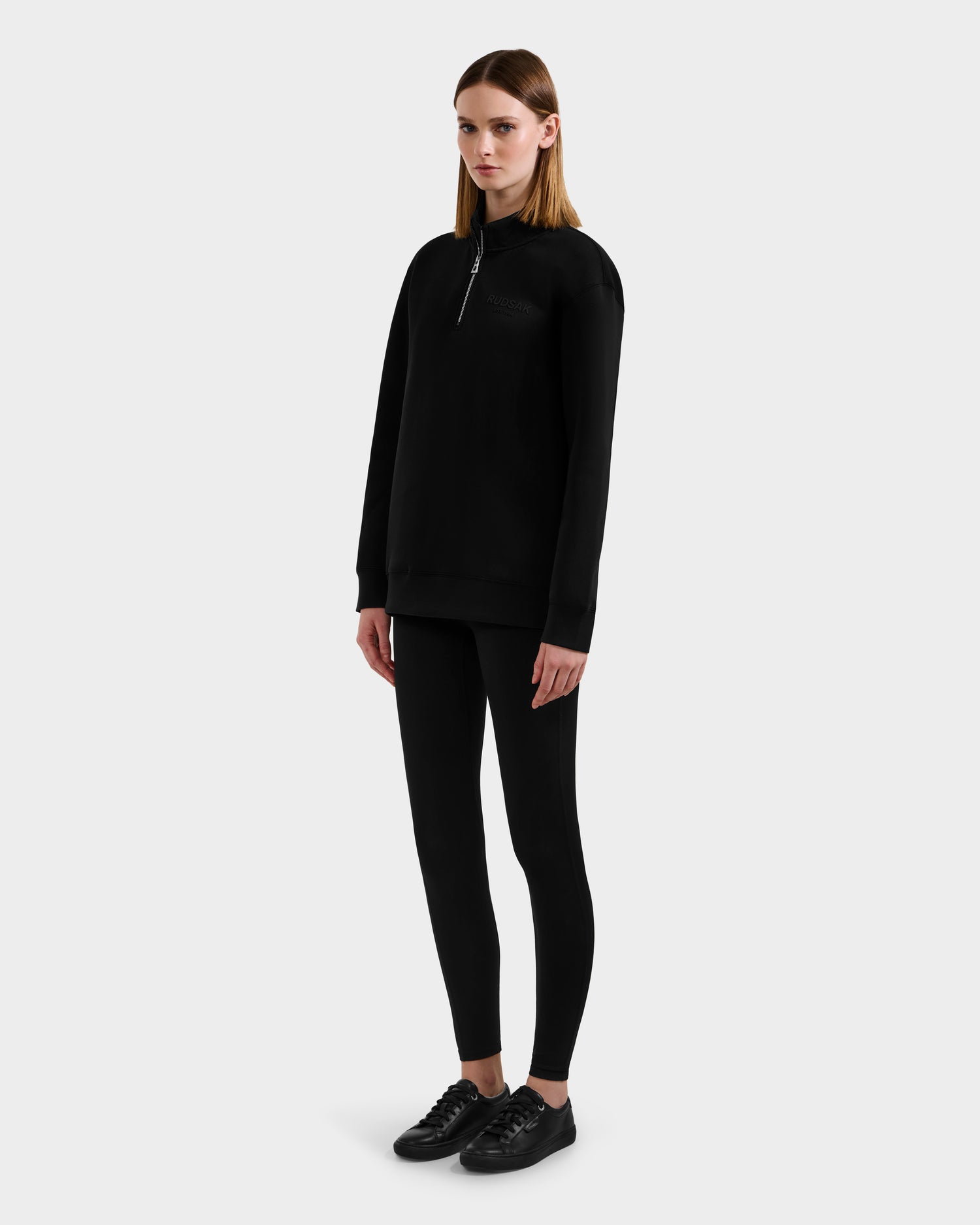 A woman faces slightly to the side, wearing the Rudsak FREMONT - 9525000 Black W zip-up sweatshirt, paired with black leggings and sneakers against a plain light gray background.