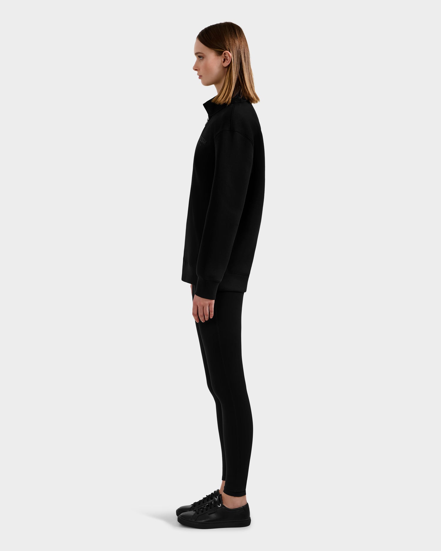 A person in profile stands against a plain background, wearing the Rudsak FREMONT - 9525000 Black W premium cotton blend sweatshirt, paired with black leggings and sneakers. Their hair is straight and shoulder-length.