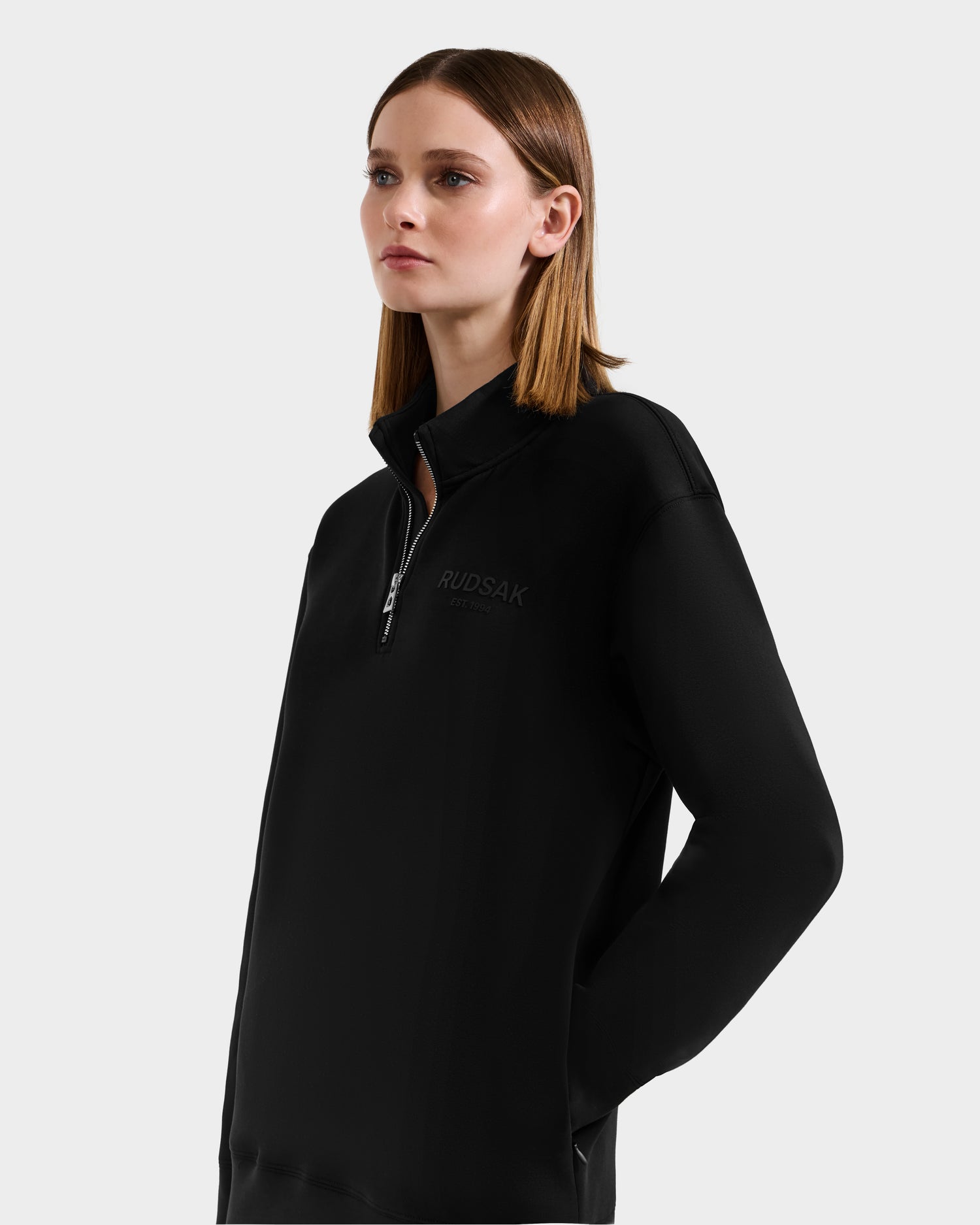 A woman with straight, shoulder-length brown hair wears the Rudsak FREMONT - 9525000 Black W mock neck cotton sweater, featuring a half-zip and chest logo, standing against a plain light grey background.