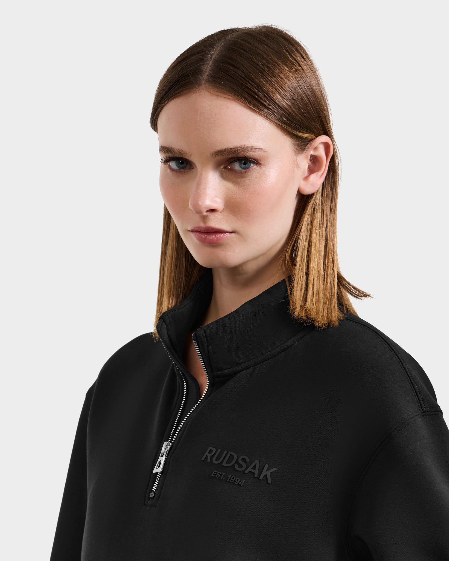 A woman with straight light brown hair wears the black Rudsak FREMONT - 9525000 W sweater, featuring a mock neck and embossed “RUDSAK EST. 1994”, standing against a plain light gray background.