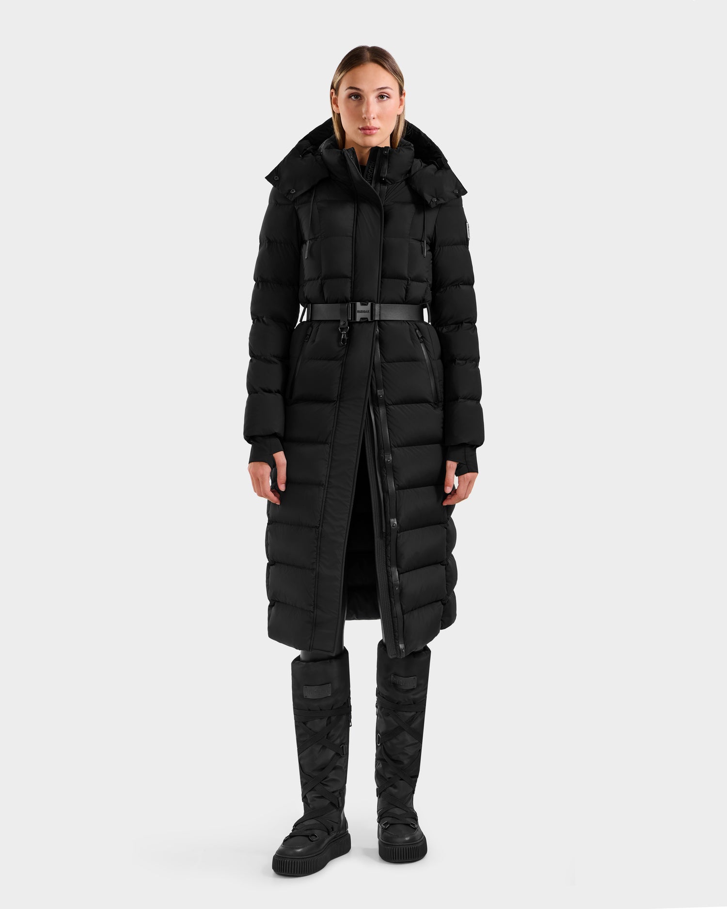 A woman faces forward in a Rudsak FRIDA M-L - L125608 Black long down puffer with hood, removable belt, black pants, and quilted boots. The coat is fully zipped with RDS-certified down. Background is plain light gray.