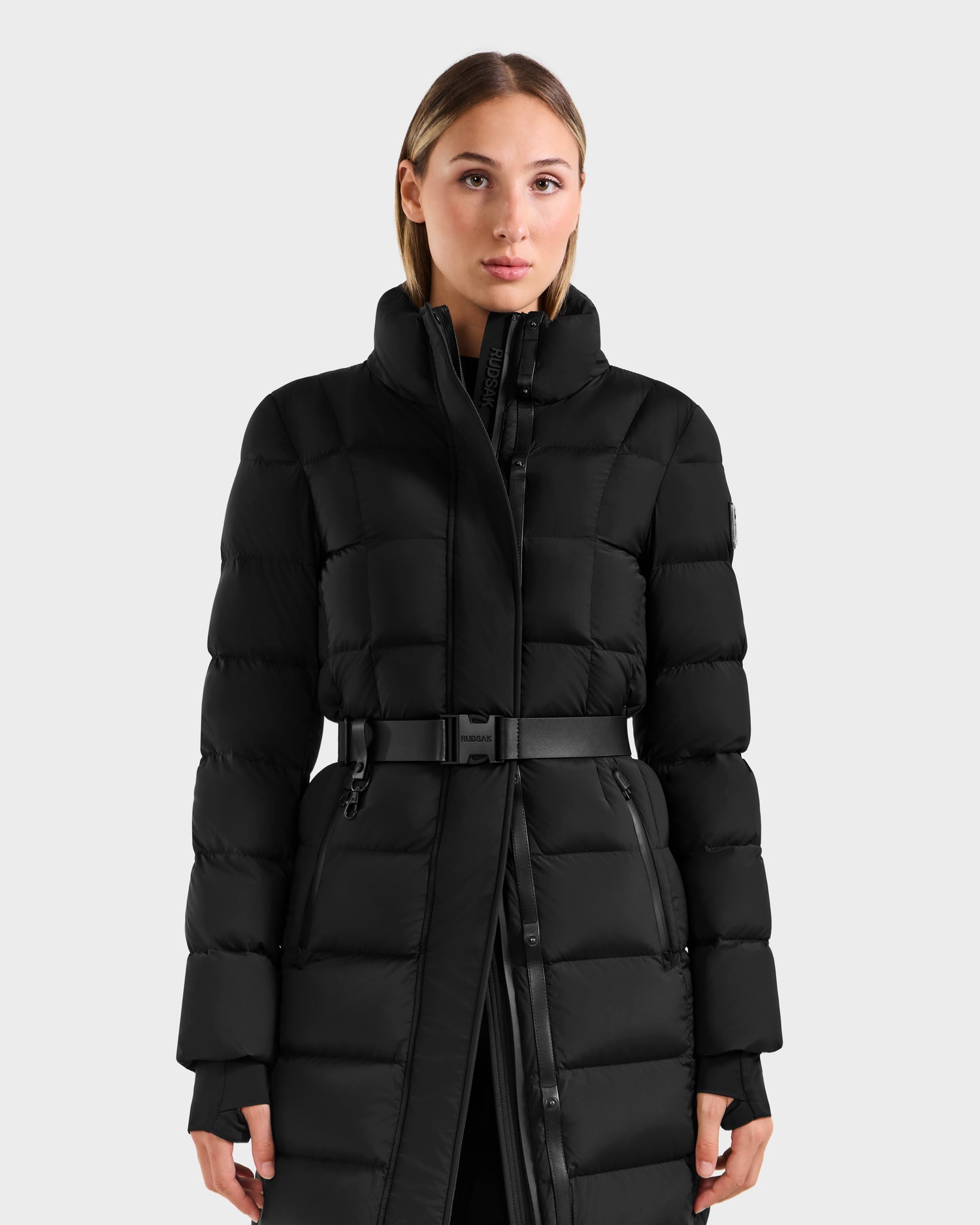A woman with straight light brown hair wears the Rudsak FRIDA M-L - L125608 Black women’s long down puffer with a high collar and removable belt, standing against a plain light gray background.