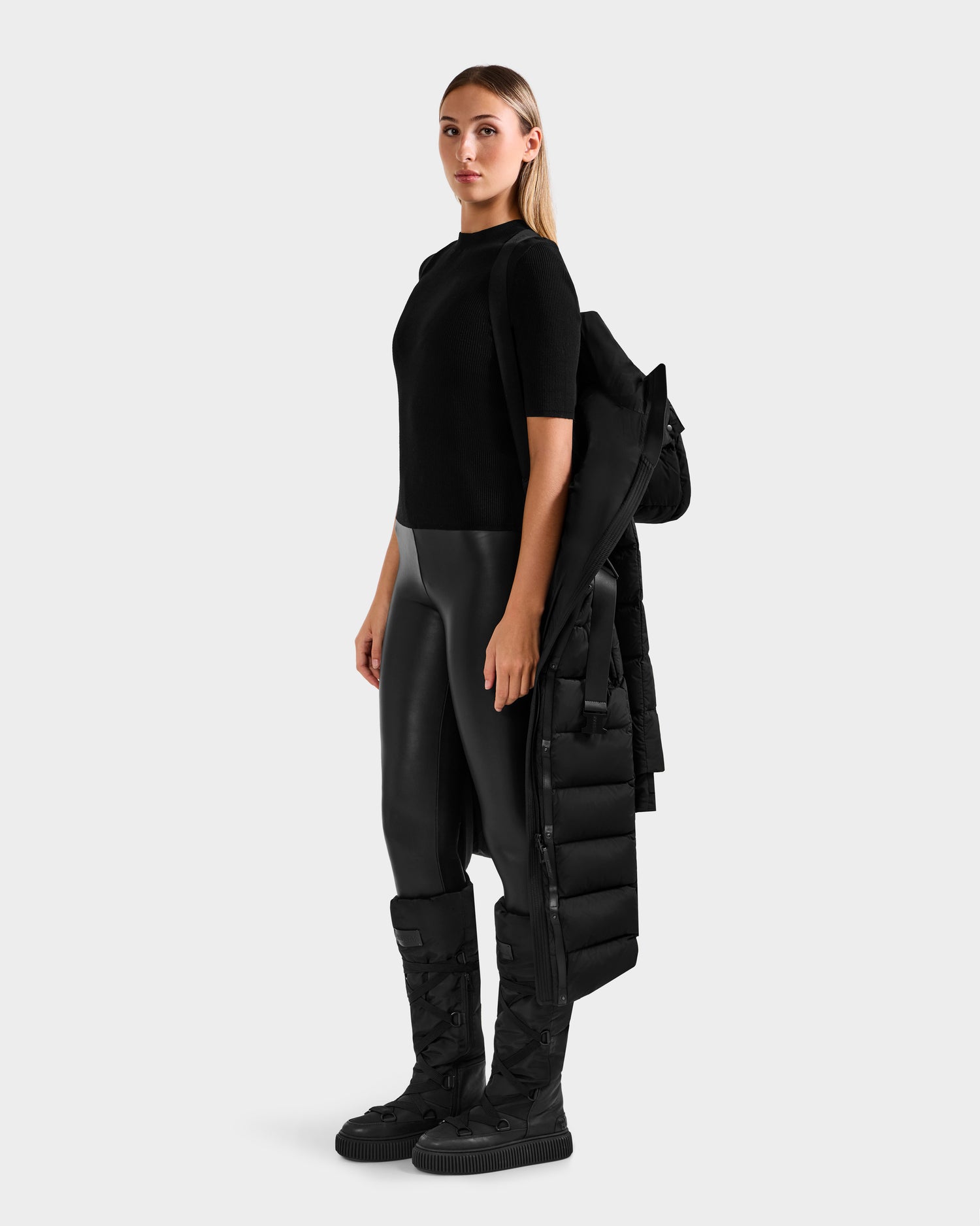 A woman stands against a plain background wearing a black short-sleeve top, faux leather pants, and tall boots. She holds the Rudsak FRIDA M-L - L125608 Black women’s long down puffer made with RDS-certified down, draped over her arm.