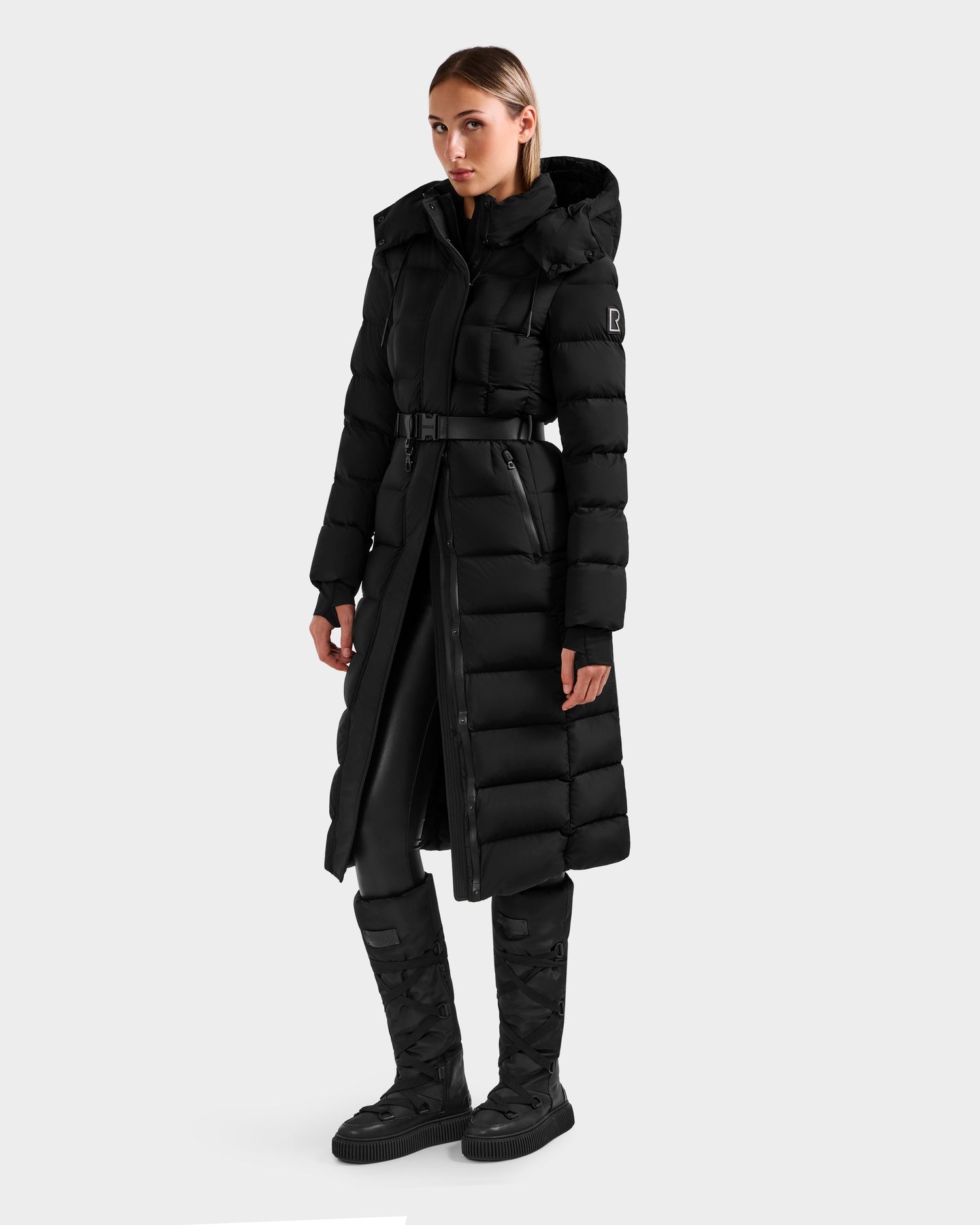 A woman wears the Rudsak FRIDA M-L - L125608 Black long down puffer with a removable belt, black pants, and matching black quilted boots, standing slightly to the side against a plain light background.