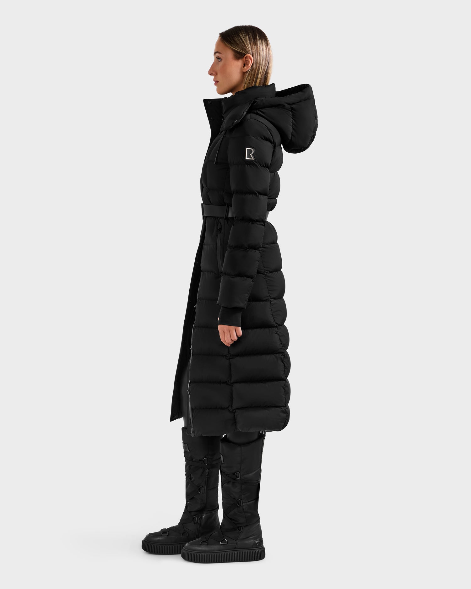 A woman stands in profile wearing the Rudsak FRIDA M-L - L125608 Black long down puffer with RDS-certified down, a removable belt, hood, and logo patch on the sleeve. She pairs it with black boots against a plain light gray background.