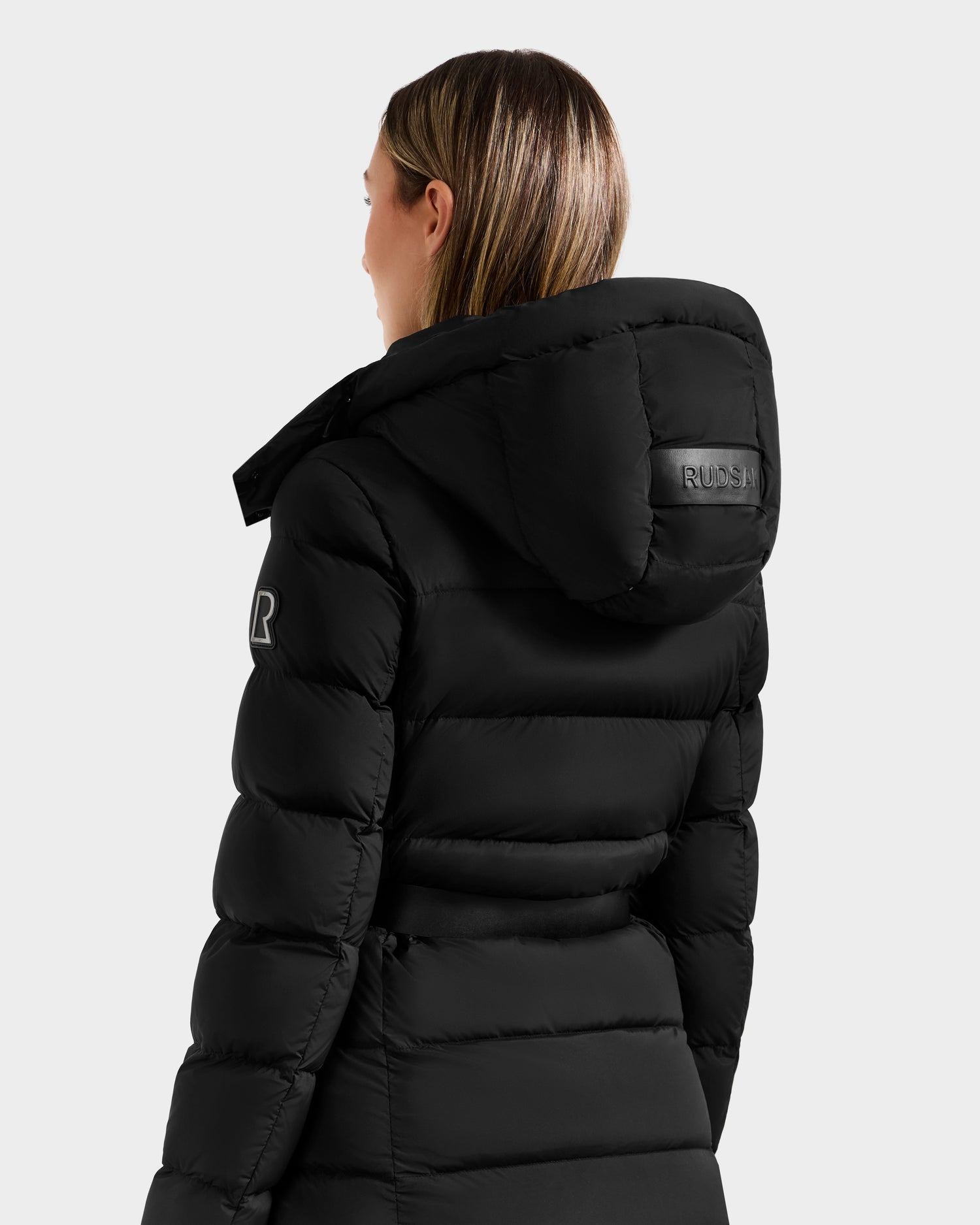 A woman with straight, light brown hair wears the Rudsak FRIDA M-L - L125608 Black women’s long down puffer jacket with a hood, showing logo patches on the hood and sleeve. She faces away against a plain light gray background.