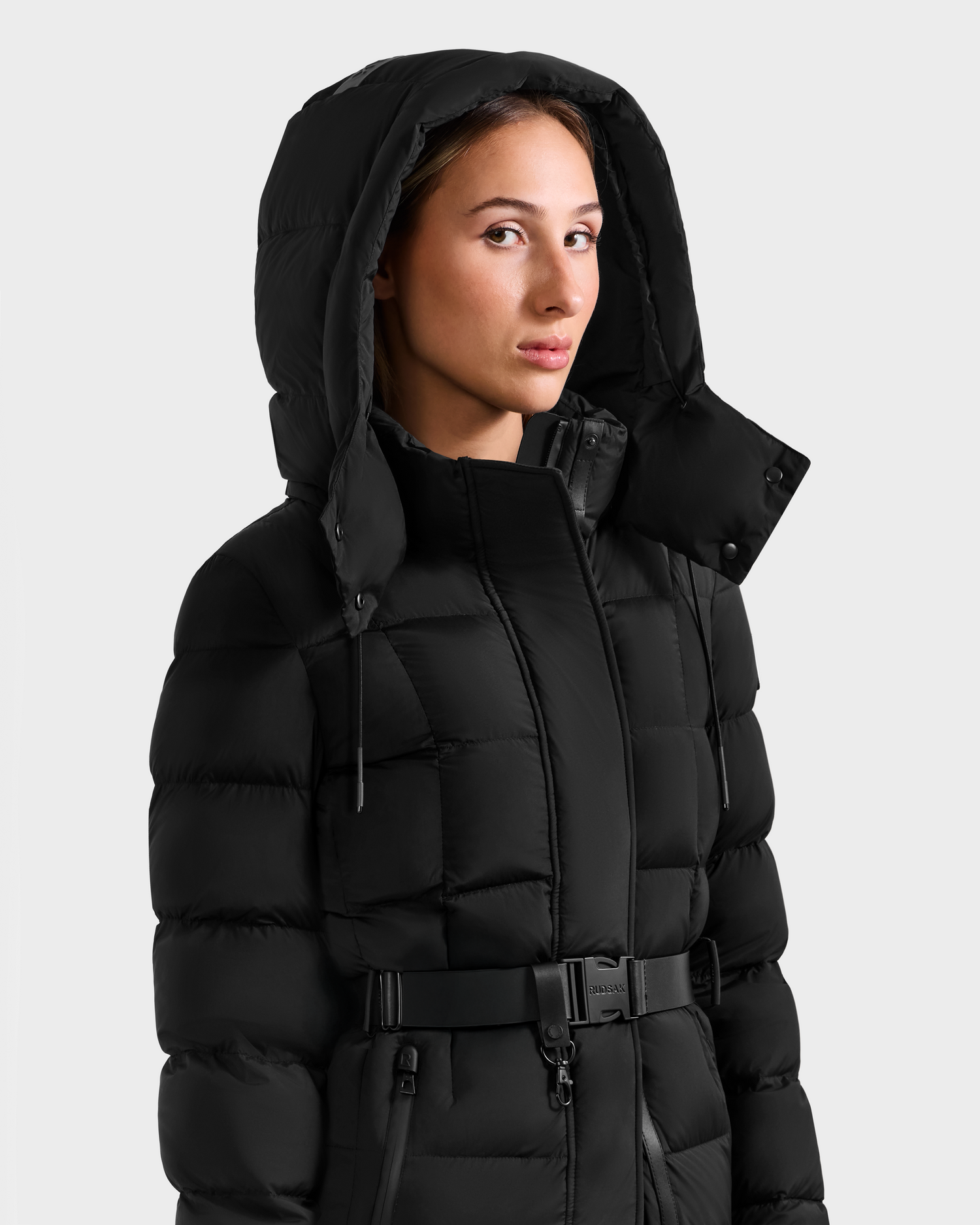 A woman wears the Rudsak FRIDA M-L - L125608 Black, a belted, hooded RDS-certified down puffer jacket, standing against a plain light gray background and looking slightly to the side.