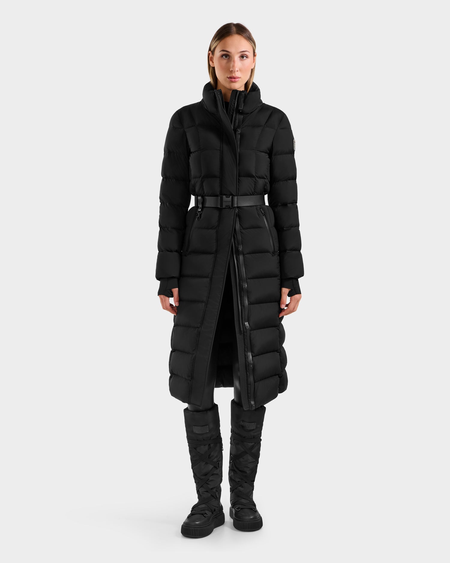 A person stands facing forward, wearing the Rudsak FRIDA M-L - L125608 Black women’s long down puffer with a removable belt and matching black puffer boots, against a plain light gray background.