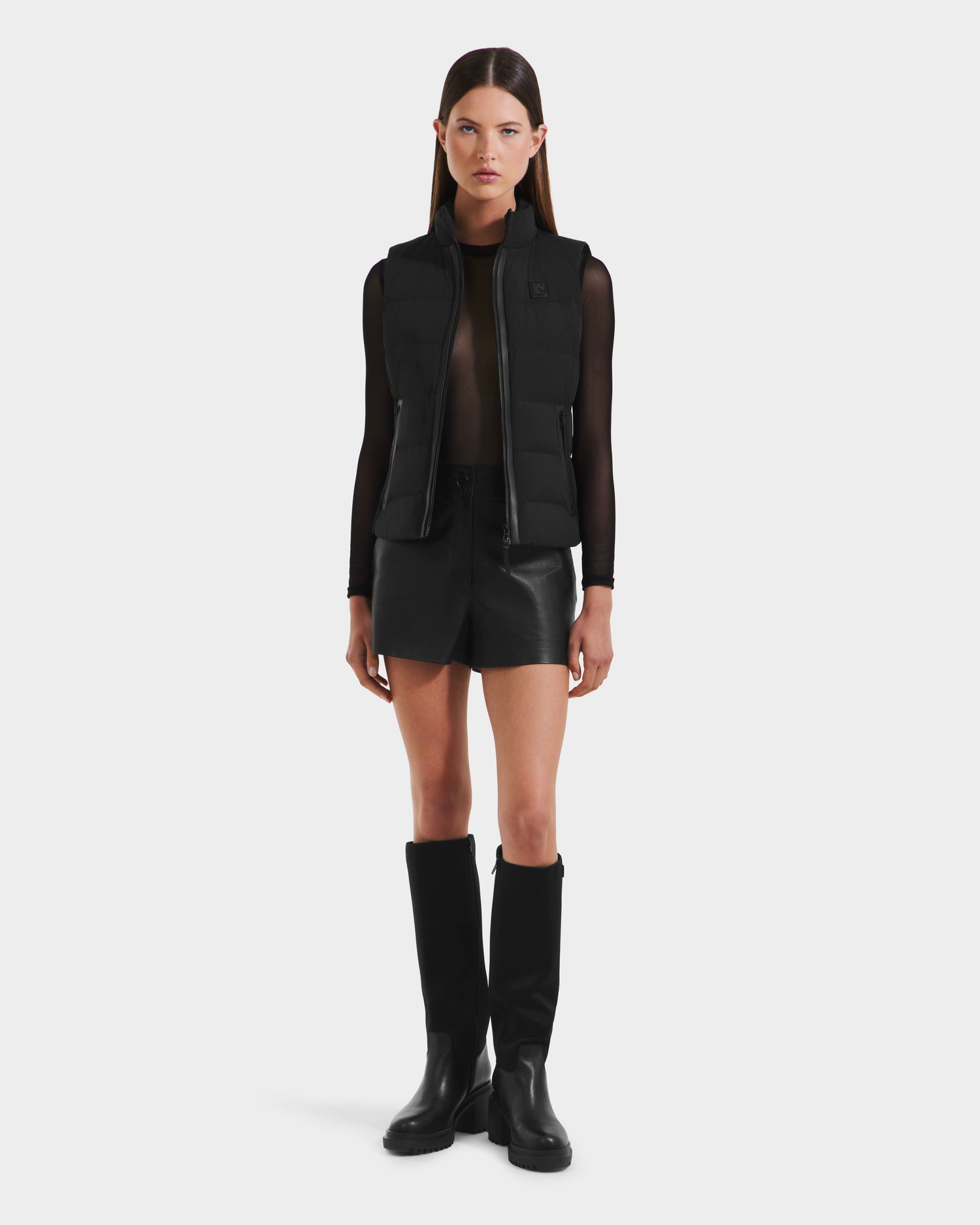 A woman faces forward against a white background wearing Rudsaks GABRIELA - 8125014 Black lightweight puffer vest with vegan leather trim, a sheer black long-sleeve top, black shorts, and knee-high black boots.