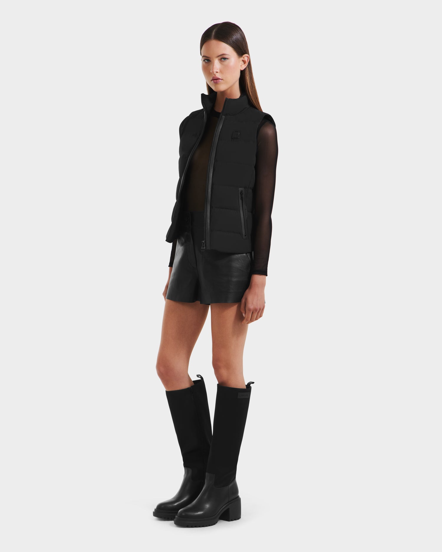 A woman stands against a plain background, wearing the Rudsak GABRIELA - 8125014 Black women’s down puffer vest over a sheer black long-sleeve top, with black leather shorts and knee-high black boots.