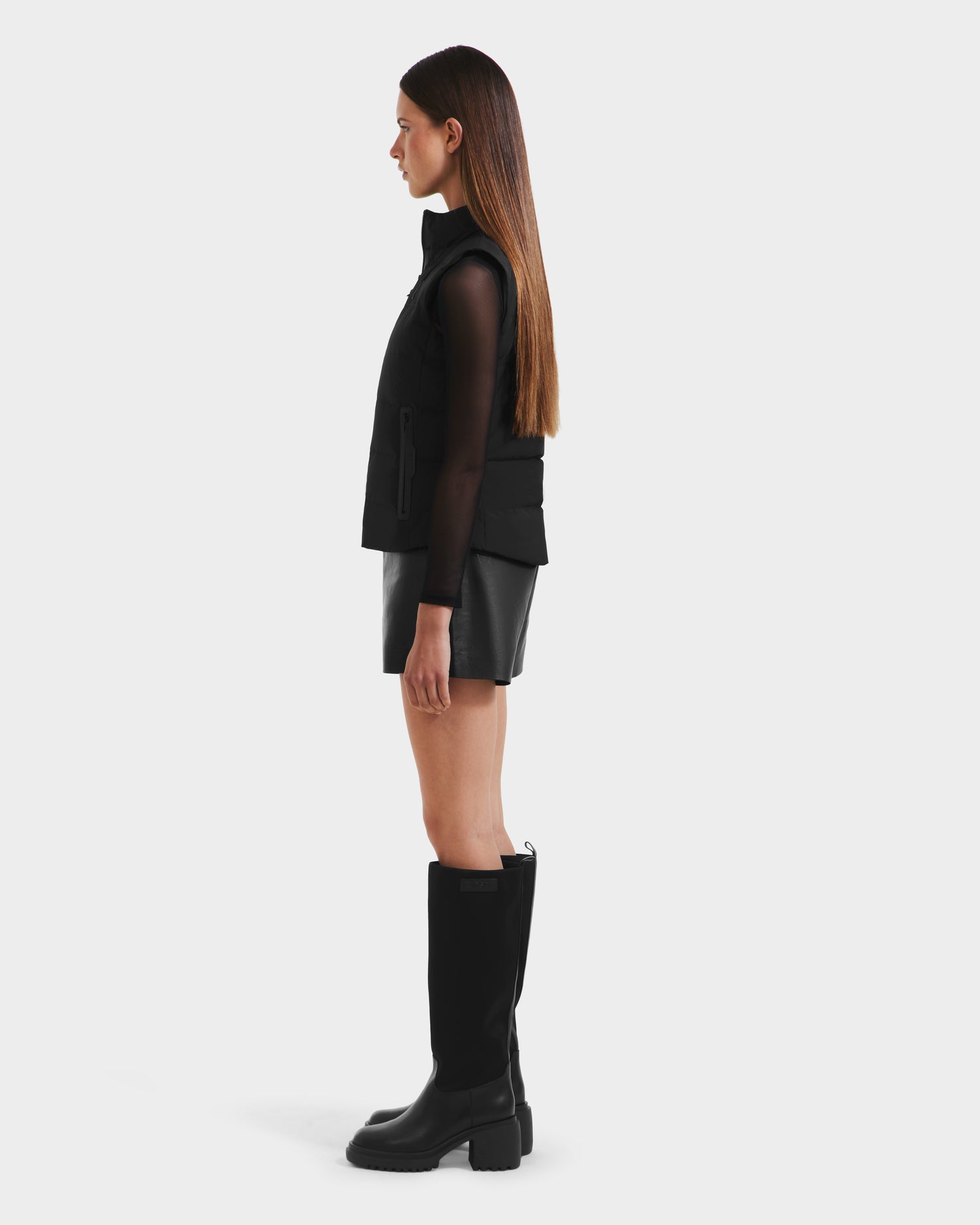 A woman stands in profile against a white background wearing the Rudsak GABRIELA - 8125014 Black down puffer vest over a black mesh long-sleeve top, black leather mini skirt, and knee-high boots; her straight hair is parted in the middle.