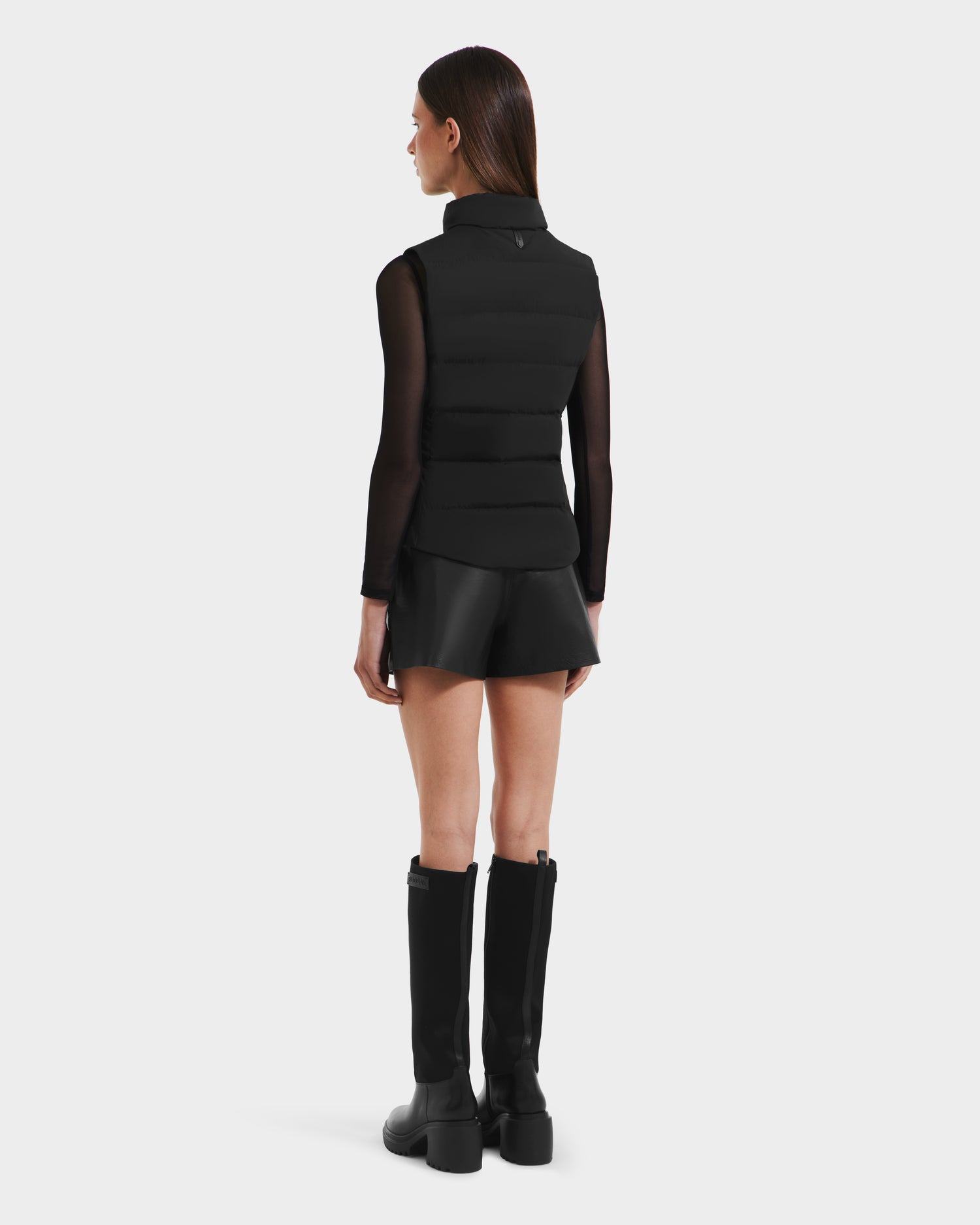 Wearing the Rudsak GABRIELA - 8125014 Black lightweight puffer vest, a woman faces away in a sheer black long-sleeve top, black leather shorts, and knee-high boots against a white background.