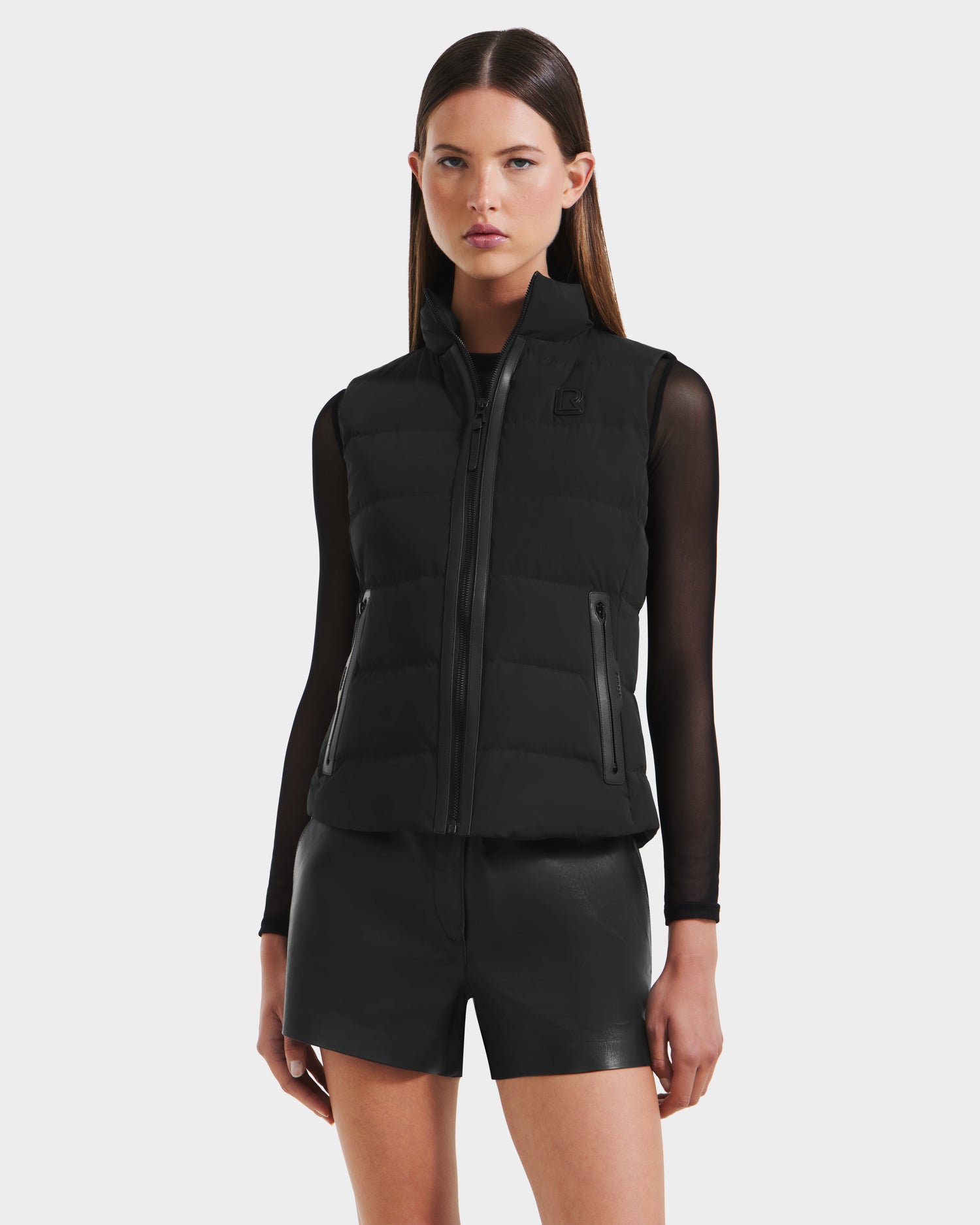 A woman faces forward against a light background, wearing the Rudsak GABRIELA - 8125014 Black lightweight puffer vest with vegan leather trim over a sheer black long-sleeve top and black leather shorts.