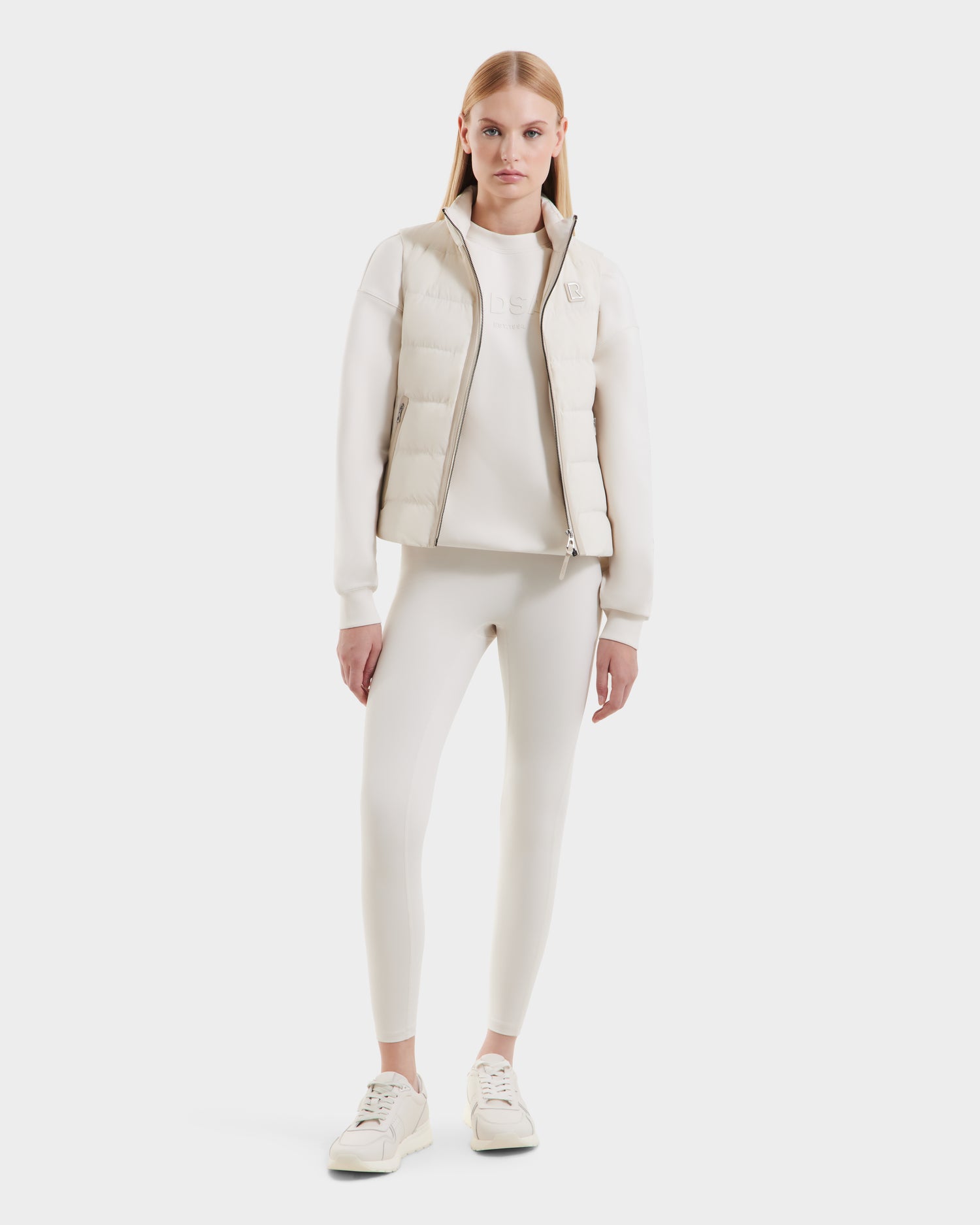 A woman faces forward against a white background, wearing the Rudsak GABRIELA - 8125014 Vanilla womens down puffer vest with vegan leather trims, plus a white long-sleeve shirt, leggings, and sneakers.