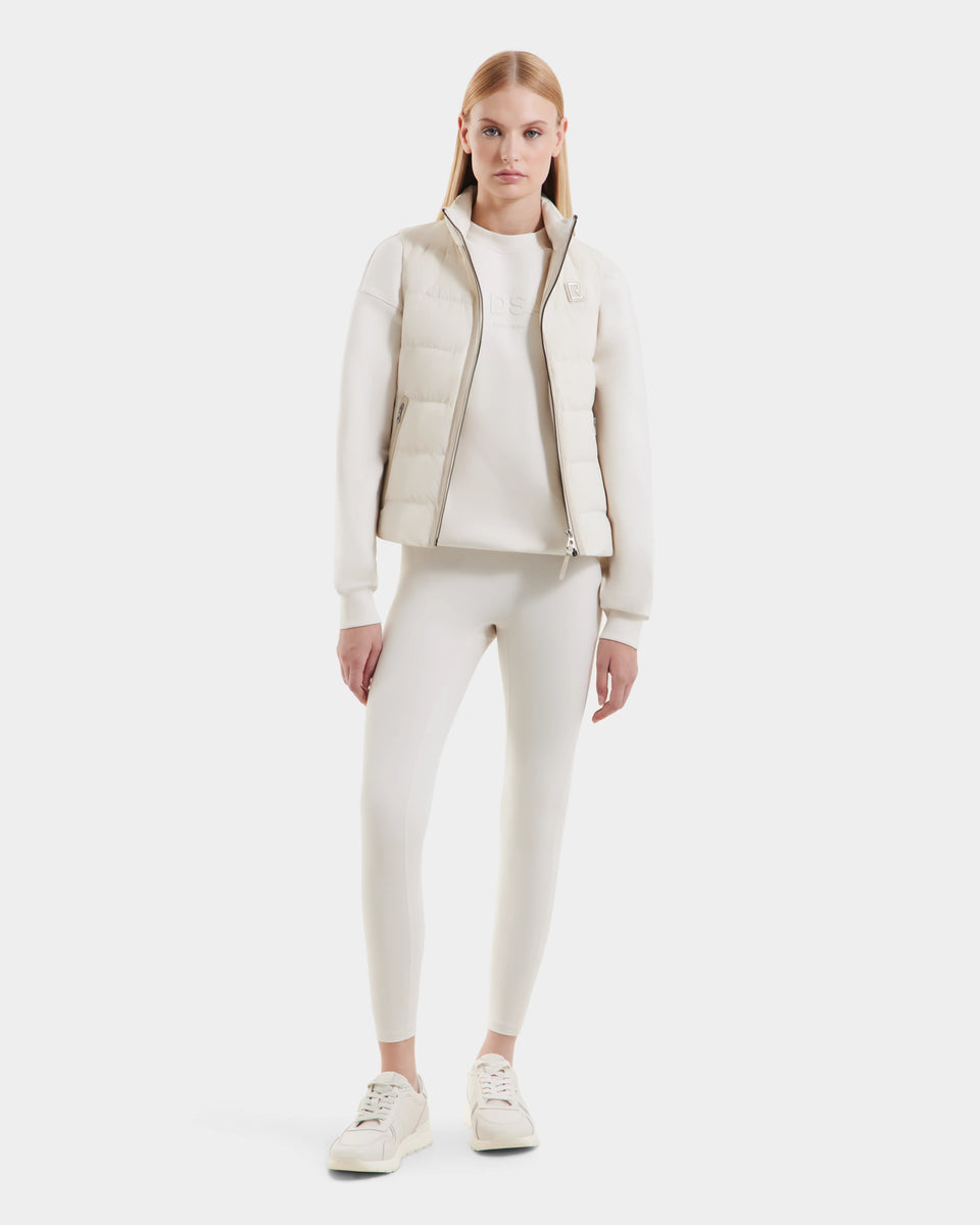 A woman faces forward against a white background, wearing the Rudsak GABRIELA - 8125014 Vanilla womens down puffer vest with vegan leather trims, plus a white long-sleeve shirt, leggings, and sneakers.