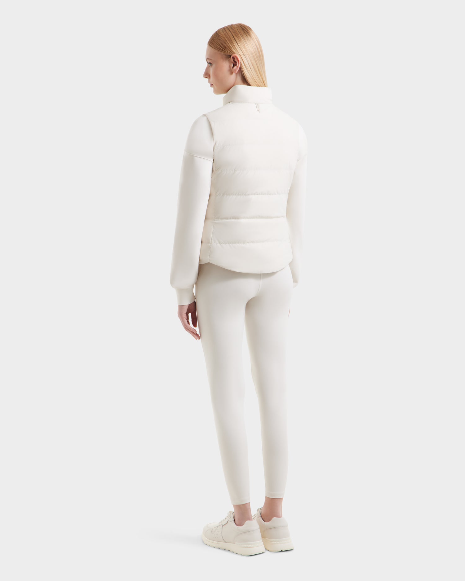 A person with straight blonde hair, dressed in Rudsak’s all-white GABRIELA - 8125014 Vanilla puffer vest with vegan leather trims, leggings, and sneakers, stands facing away against a plain light background.