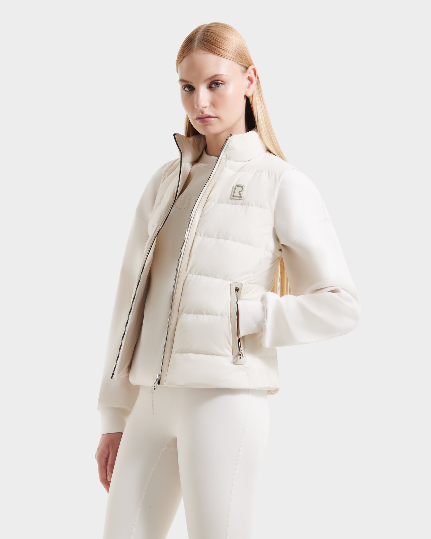 A woman with long straight blonde hair models the Rudsak GABRIELA - 8125014 Vanilla: an all-white zip-up puffer jacket with vegan leather trim and a subtle chest logo, against a plain light background.