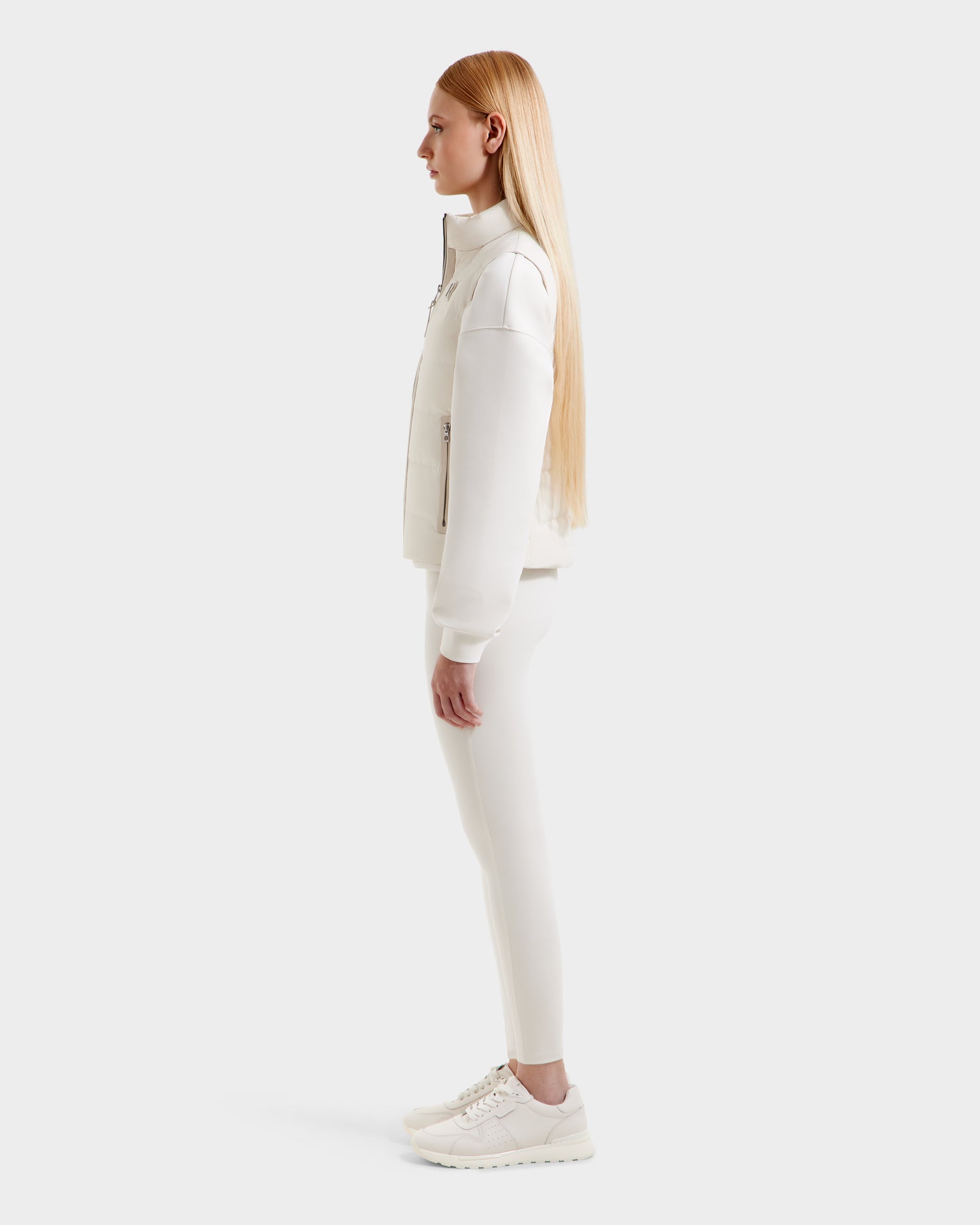 A woman with long straight blonde hair stands in profile against a plain white background, wearing the Rudsak GABRIELA - 8125014 Vanilla lightweight down puffer vest with vegan leather trims, paired with leggings and sneakers.