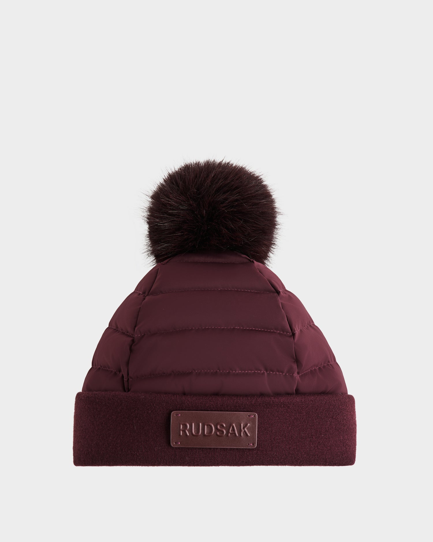 The Rudsak GALA - 8424514 Bordeaux is a maroon quilted winter hat with a large matching faux fur pom pom, a folded cuff, and a RUDSAK label on the front, shown against a plain white background.
