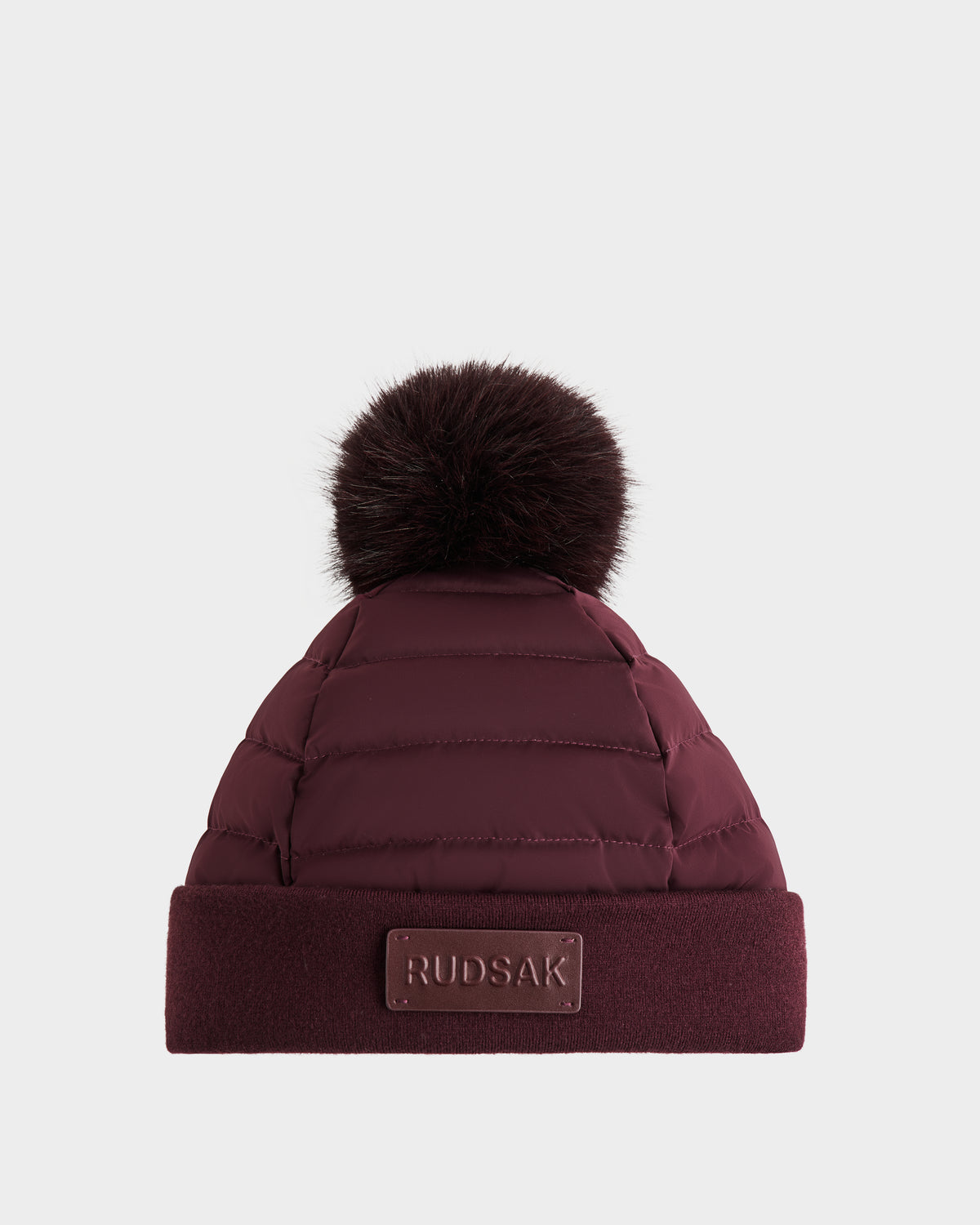 The Rudsak GALA - 8424514 Bordeaux is a maroon quilted winter hat with a large matching faux fur pom pom, a folded cuff, and a RUDSAK label on the front, shown against a plain white background.
