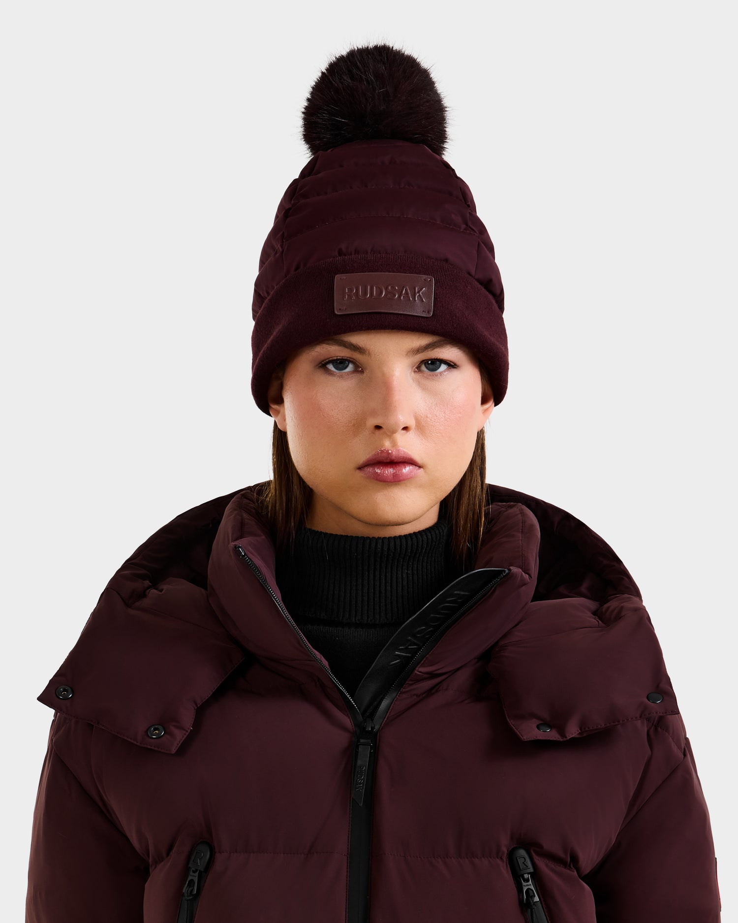 A woman in the Rudsak GALA - 8424514 Bordeaux quilted down puffer hat with faux fur pom pom and matching jacket stands against a light background, facing the camera with a neutral expression.