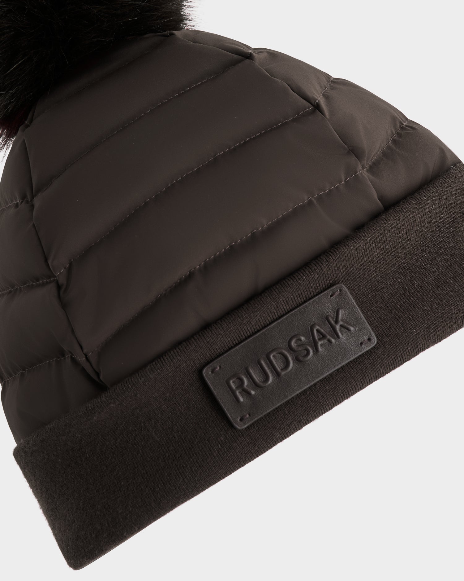 A close-up of the Rudsak GALA - 8424514 Espresso women's quilted down puffer hat, featuring a folded brim, visible brand patch, and a fluffy faux fur pom pom for stylish warmth.