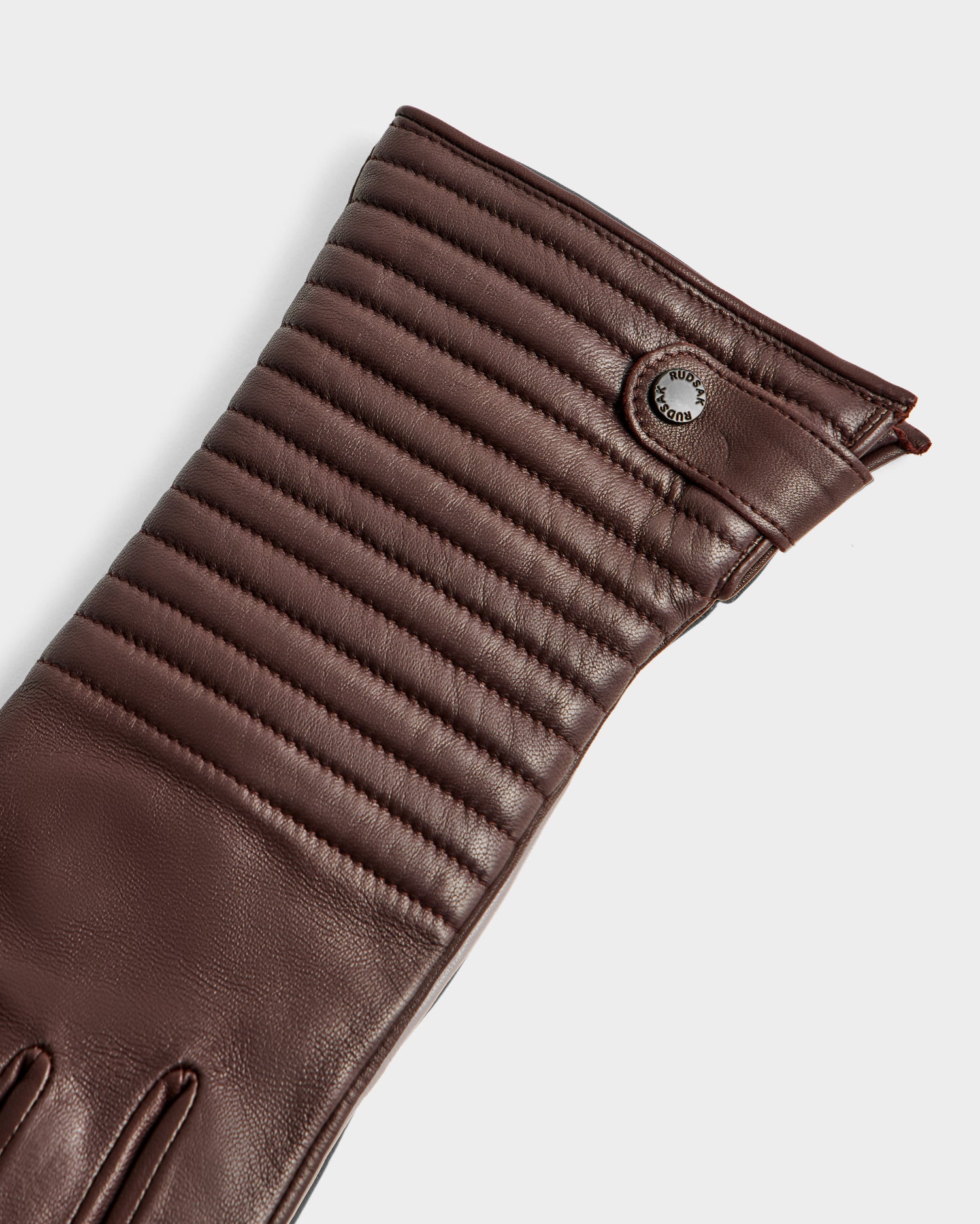 A close-up of the GEMMA - 8425008 Espresso brown leather glove by Rudsak, showcasing horizontal quilted stitching on the cuff and a small round metallic button detail near the wrist—this season’s essential winter accessory.